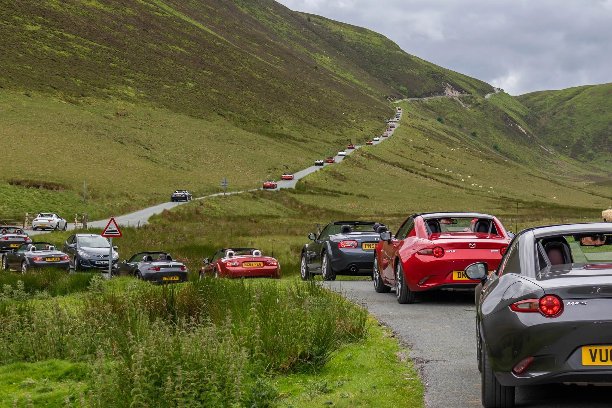 A long line of cars parked on a narrow mountain road surrounded by green hills and grass, with some cars on the road and others on the grass.