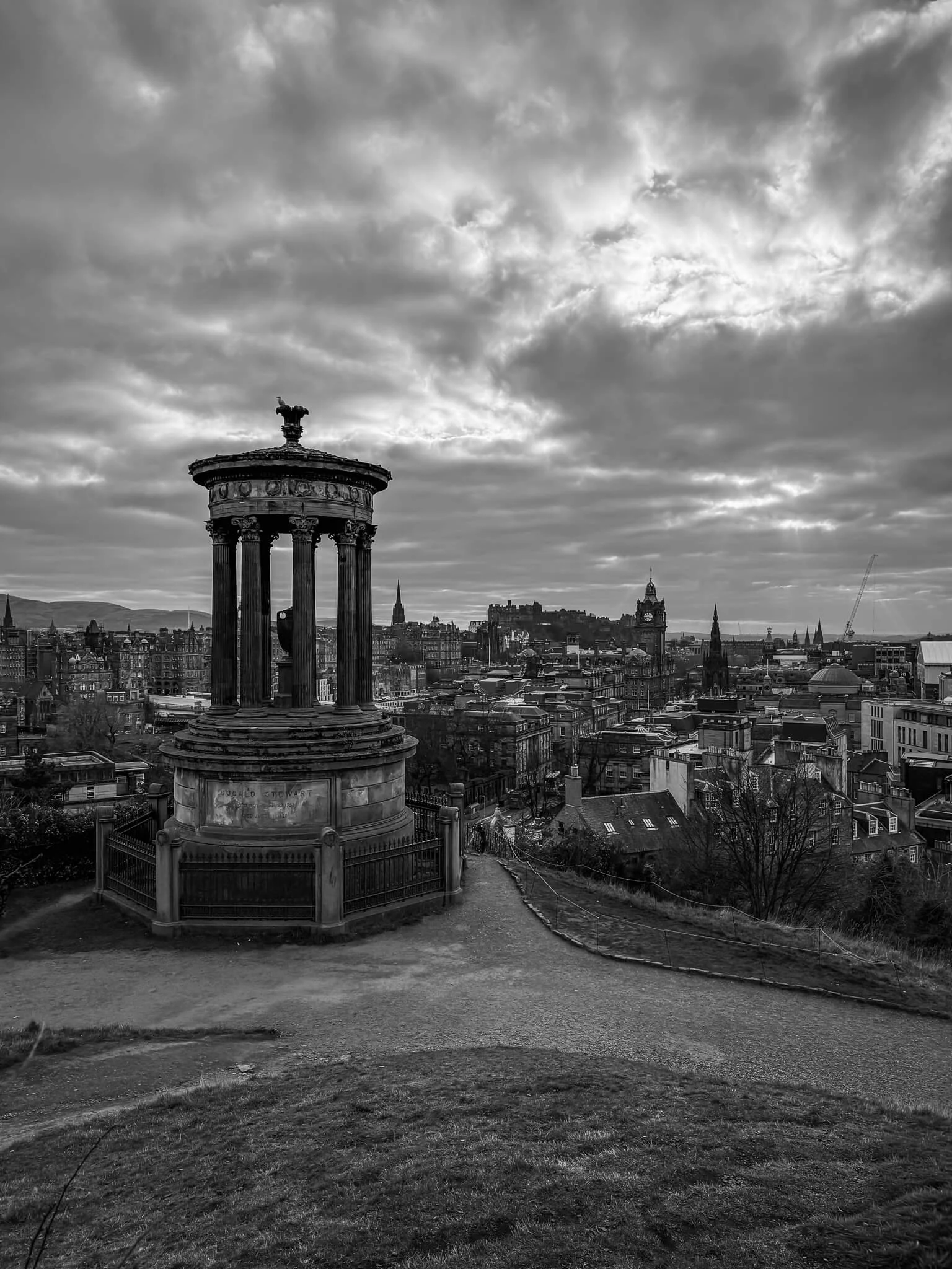 Black and white photo of a cityscape view from a hill, showing historic architecture, with a prominent circular monument with pillars in the foreground, under a cloudy sky.