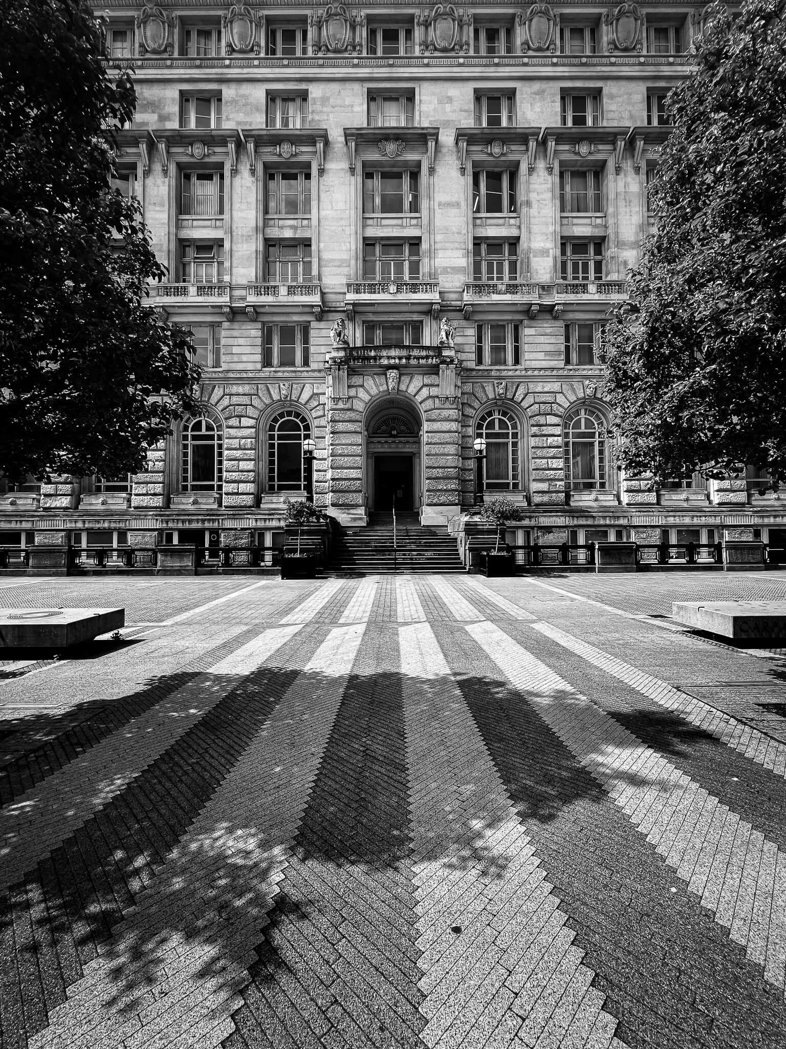 Black and white photo of a grand, historic building with arched windows, statues, and ornate architectural details, with trees on either side and a patterned brick crosswalk in the foreground.
