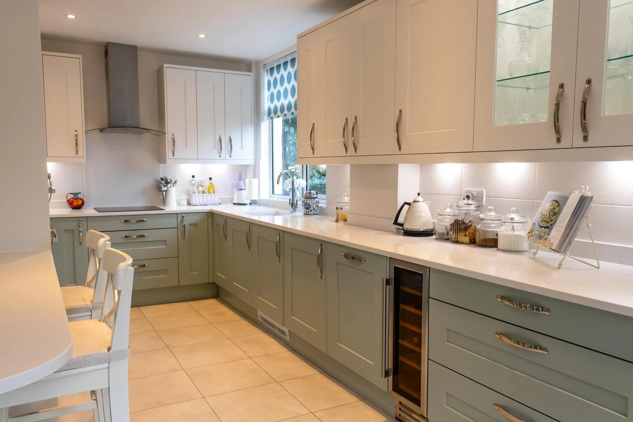Modern kitchen with beige cabinets, a white countertop, and a window with a blue and white patterned blind. There are glass jars, a toaster, and a cookbook on a stand on the counter. A small wine fridge is built into the lower cabinets, and a stainless steel range hood is above the stove.