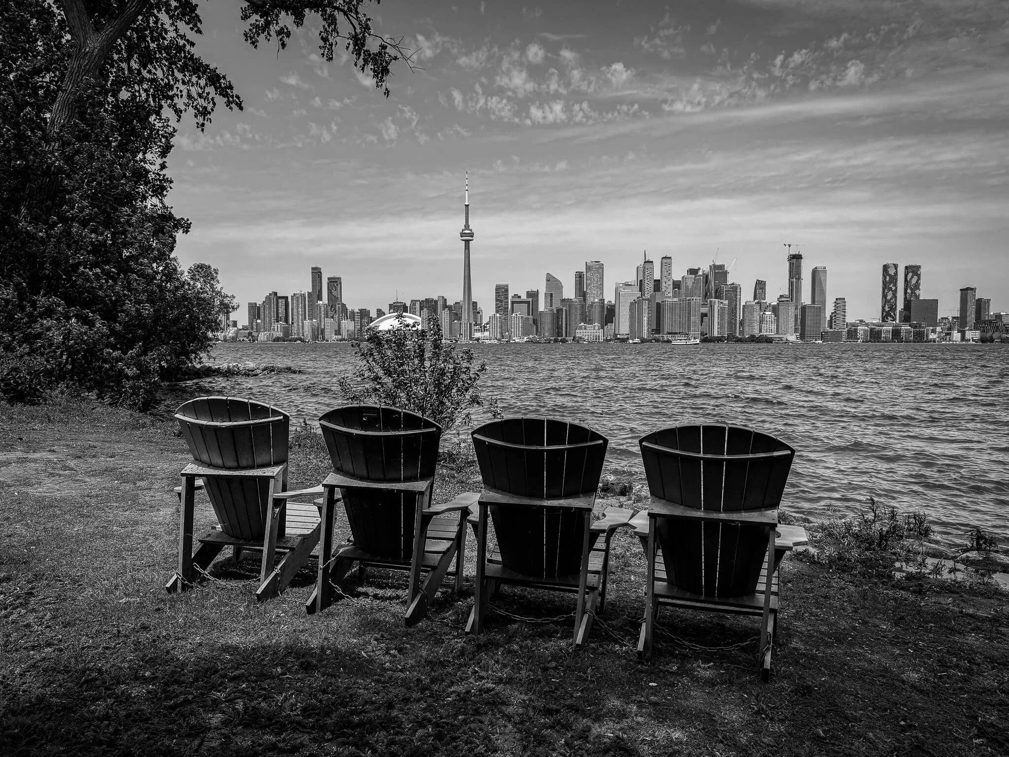 Four Adirondack chairs facing a water body with a city skyline featuring CN Tower in the background, under a partly cloudy sky.