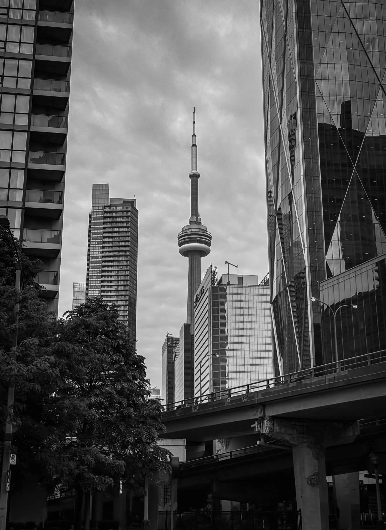 Black and white photo of downtown Toronto with the CN Tower in the center, surrounded by modern high-rise buildings and a street with a bridge.