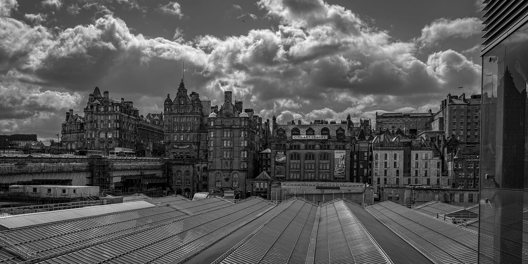 Black and white photo of city skyline with historic buildings under cloudy sky and modern rooftop structure in foreground.