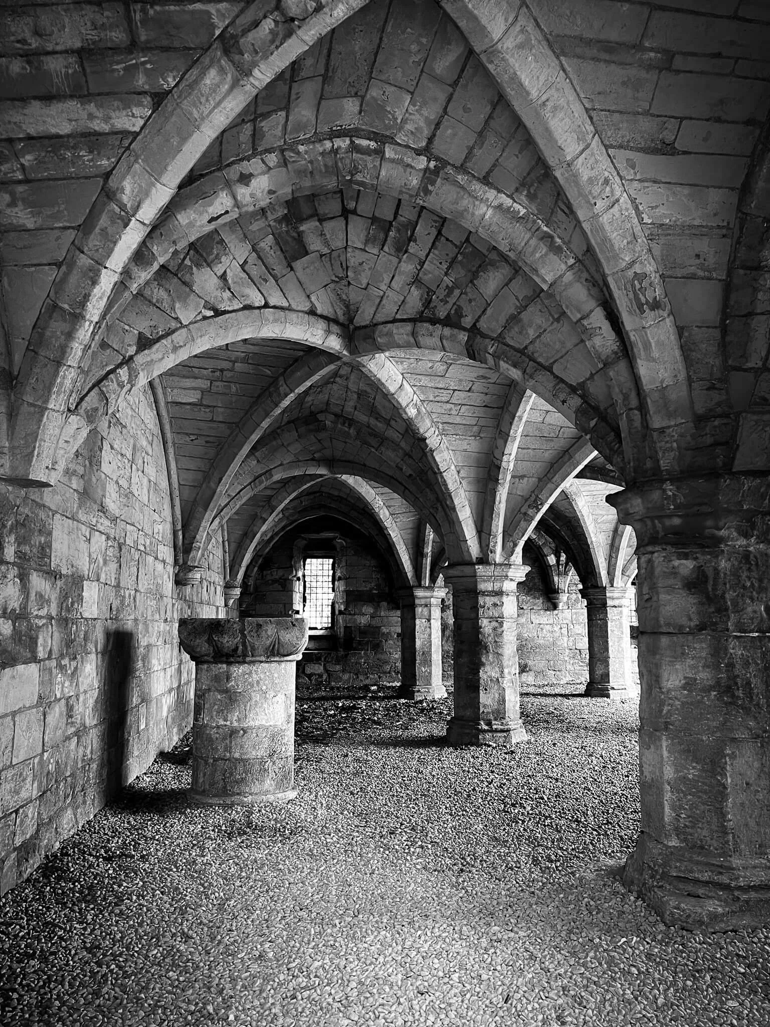 Black and white photo of stone Gothic arches and columns in an ancient hall or cloister, with a small barred window at the end.