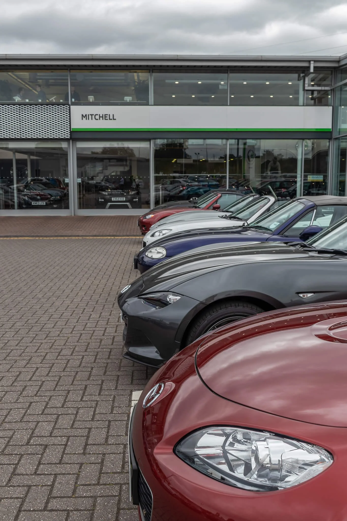 Line of parked cars outside a Volkswagen dealership with the sign 'Mitchell' on the building.