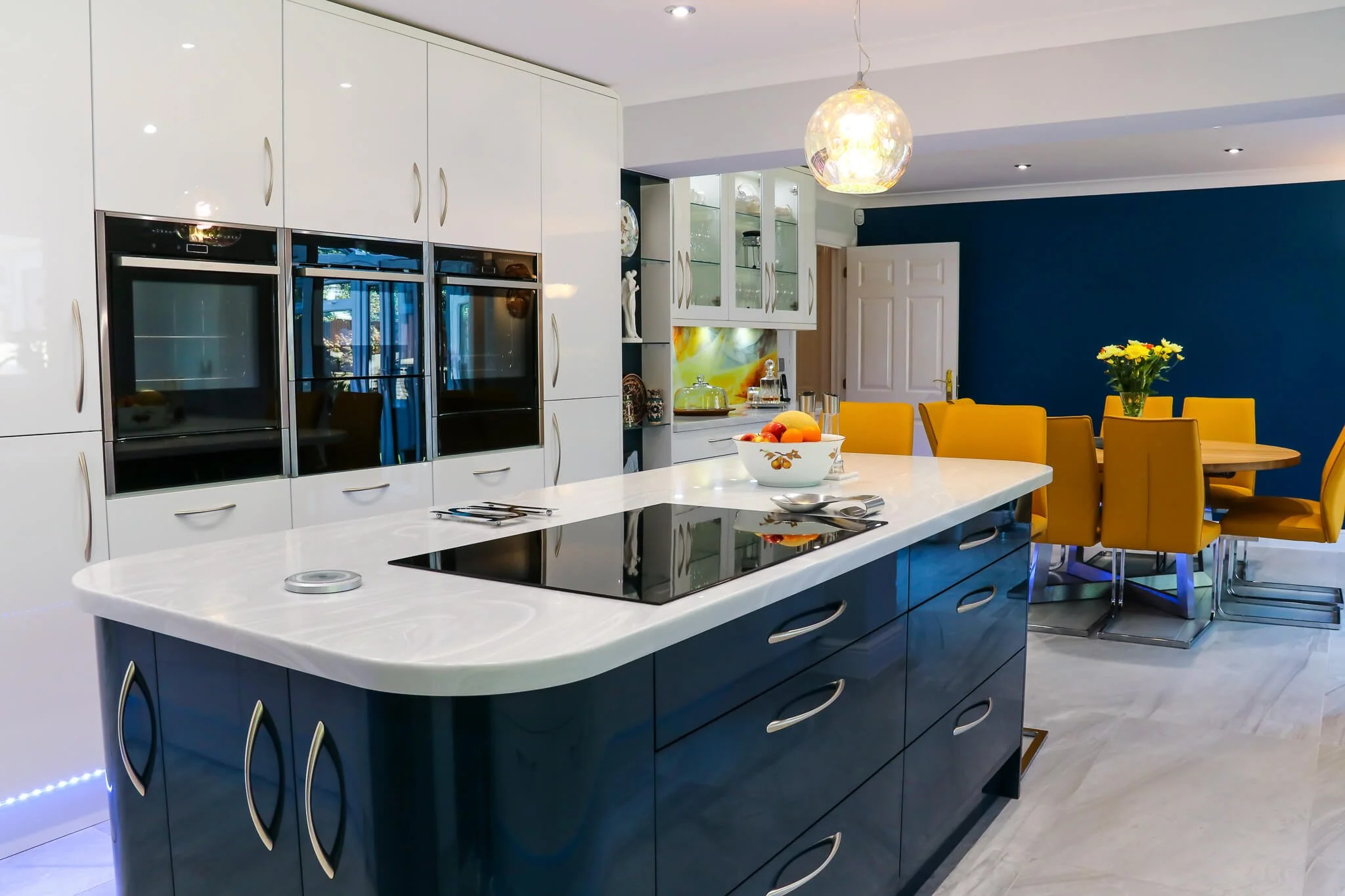 Modern kitchen with a white and blue color scheme, featuring a large central island with a white countertop, yellow dining chairs around a round table, and built-in ovens along the wall.