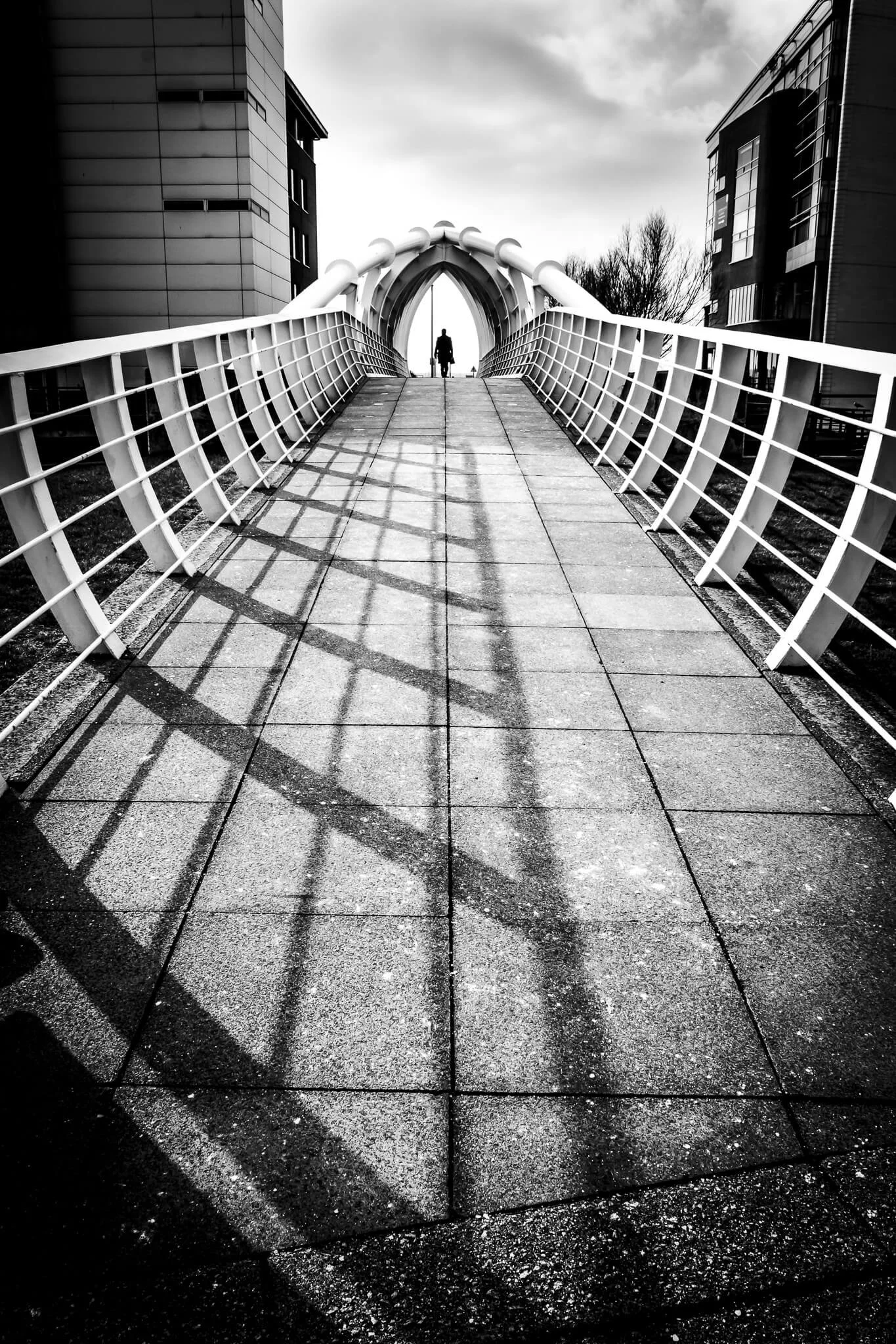 Black and white photo of a modern pedestrian bridge with a person walking in the distance, flanked by buildings on either side.