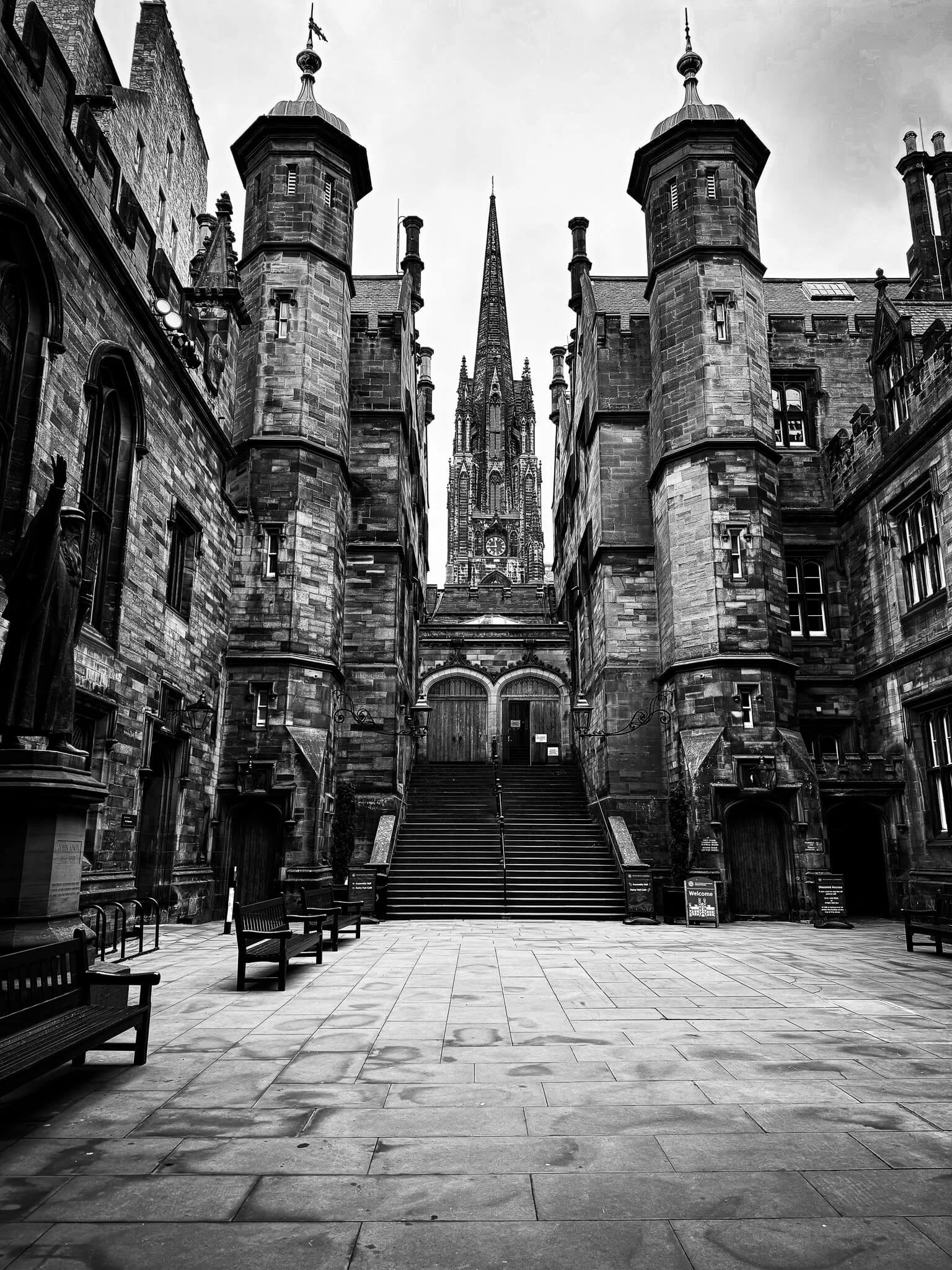 Black and white photo of a historic Gothic-style church with tall spire and castle-like stone walls, viewed from an empty courtyard with benches.