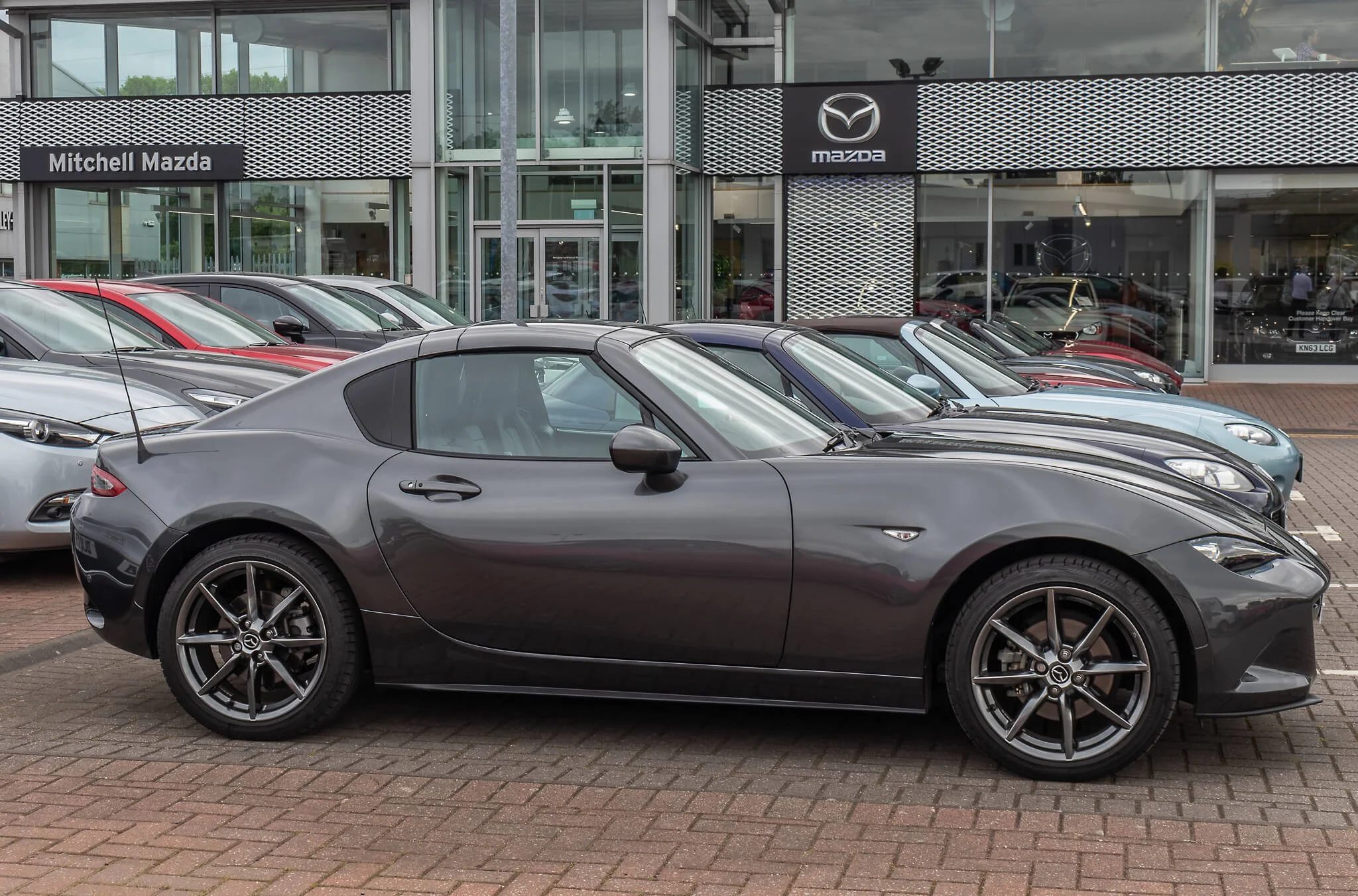 Gray Mazda convertible parked in front of a Mazda dealership, with other cars parked nearby.