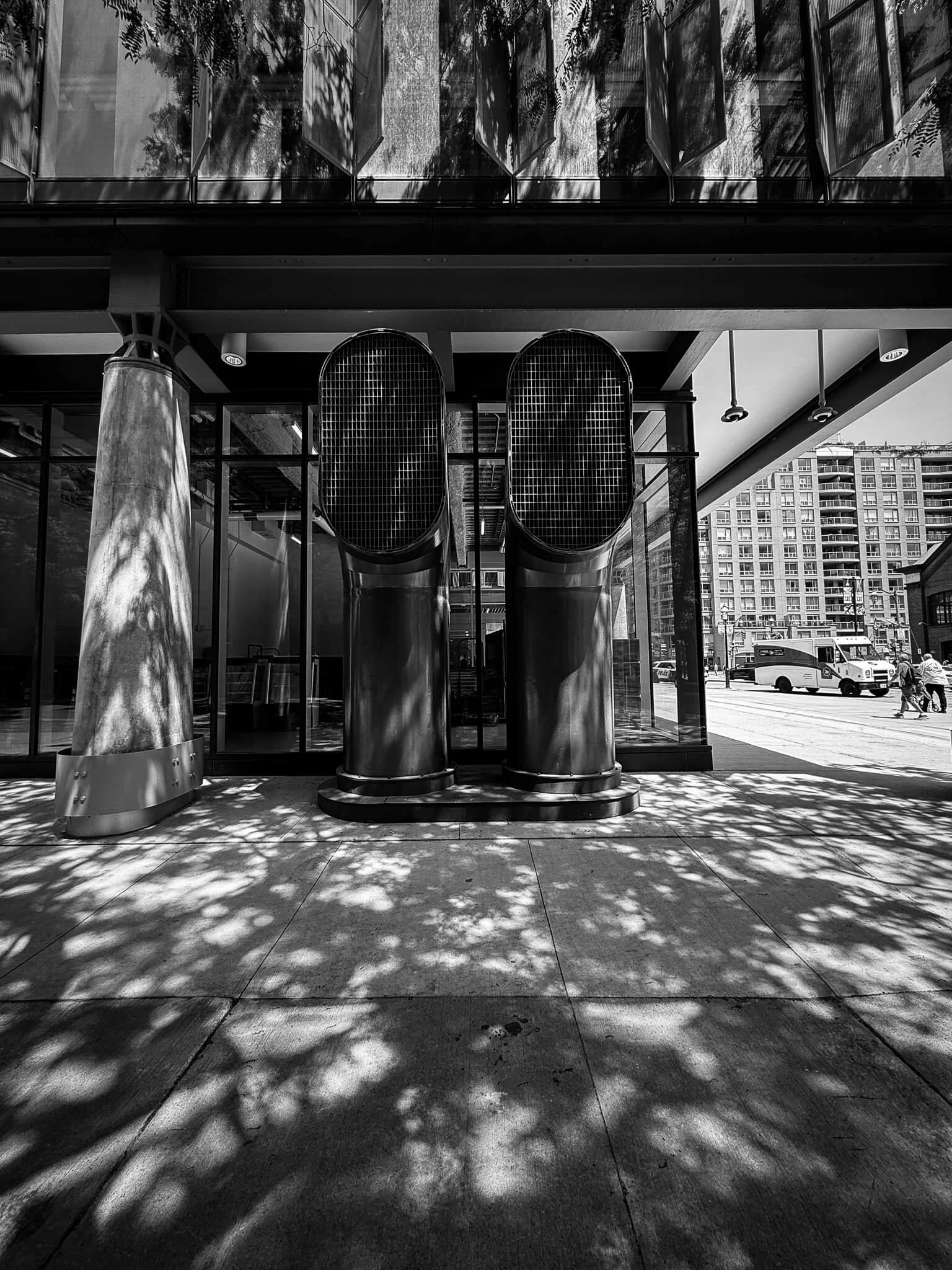 Black and white photo of two large vertical air conditioning units or vents outside a building, with shadows of trees on the sidewalk and building.