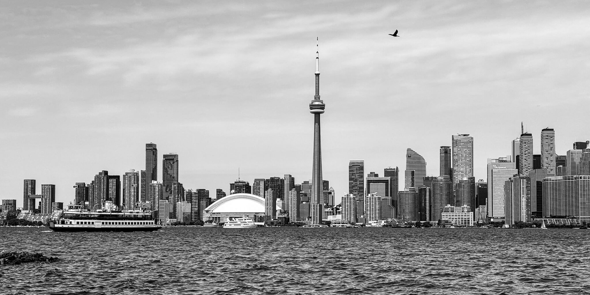 Black and white image of the Toronto skyline viewed from across Lake Ontario, featuring the CN Tower prominently in the center with surrounding skyscrapers and a boat on the water.