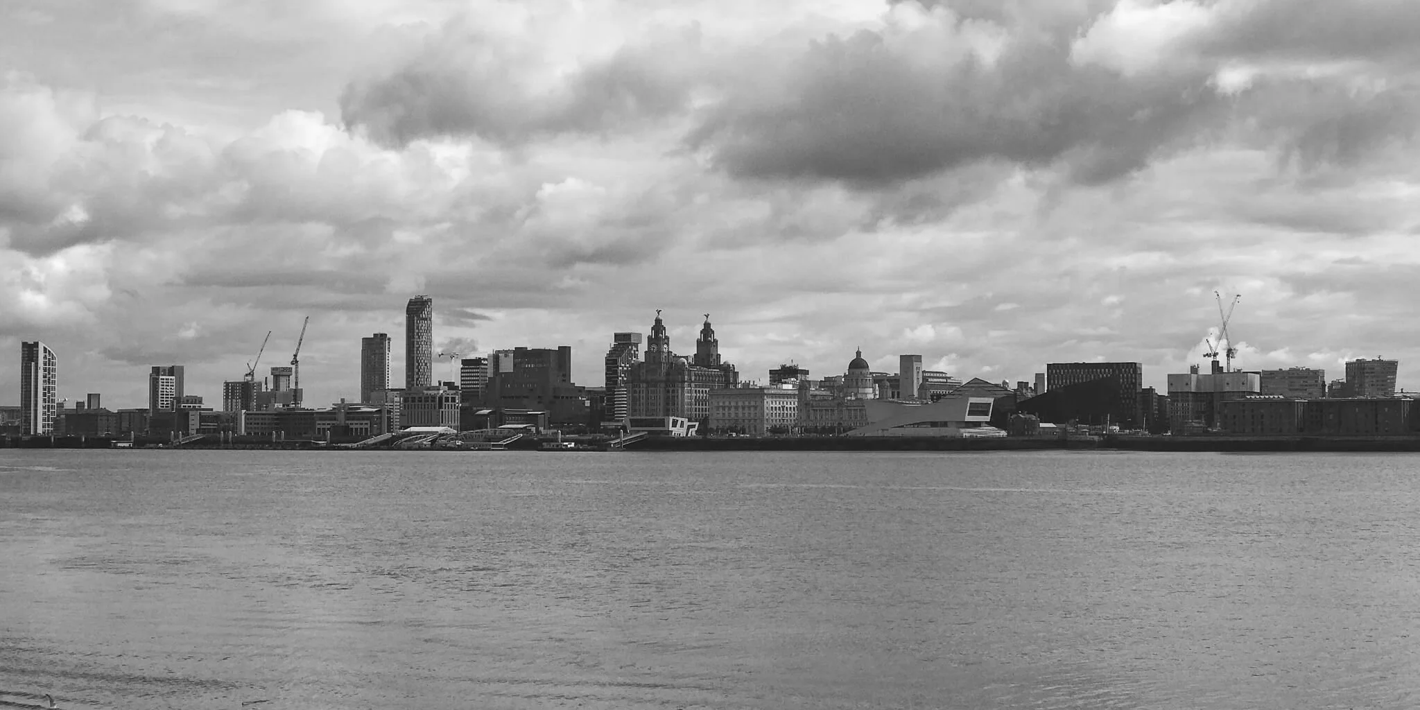 Black and white photo of a city skyline across a body of water with cloudy sky overhead.