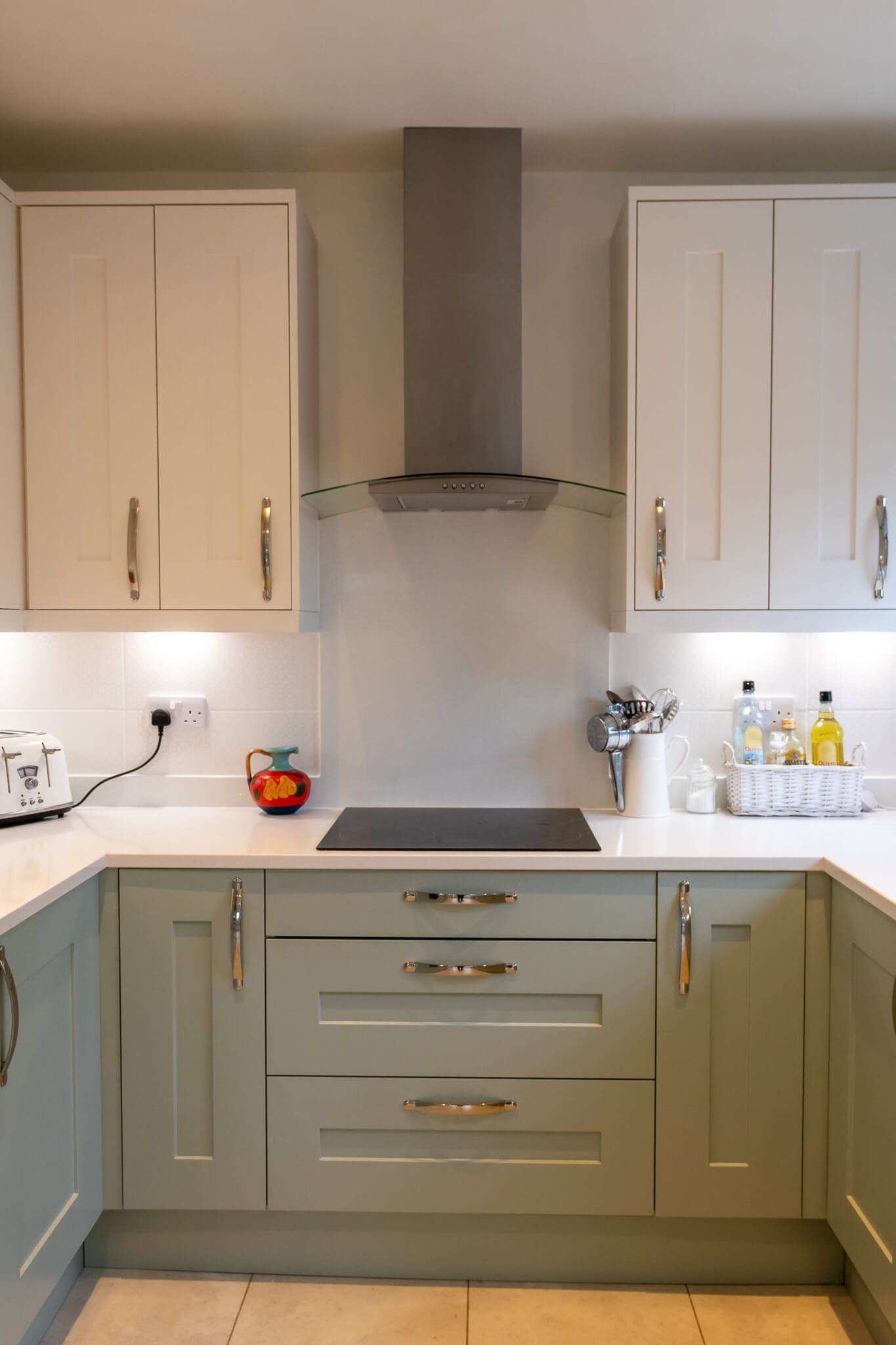 Modern kitchen with beige cabinets, a black cooktop, a range hood, and various small appliances and bottles on the countertop.