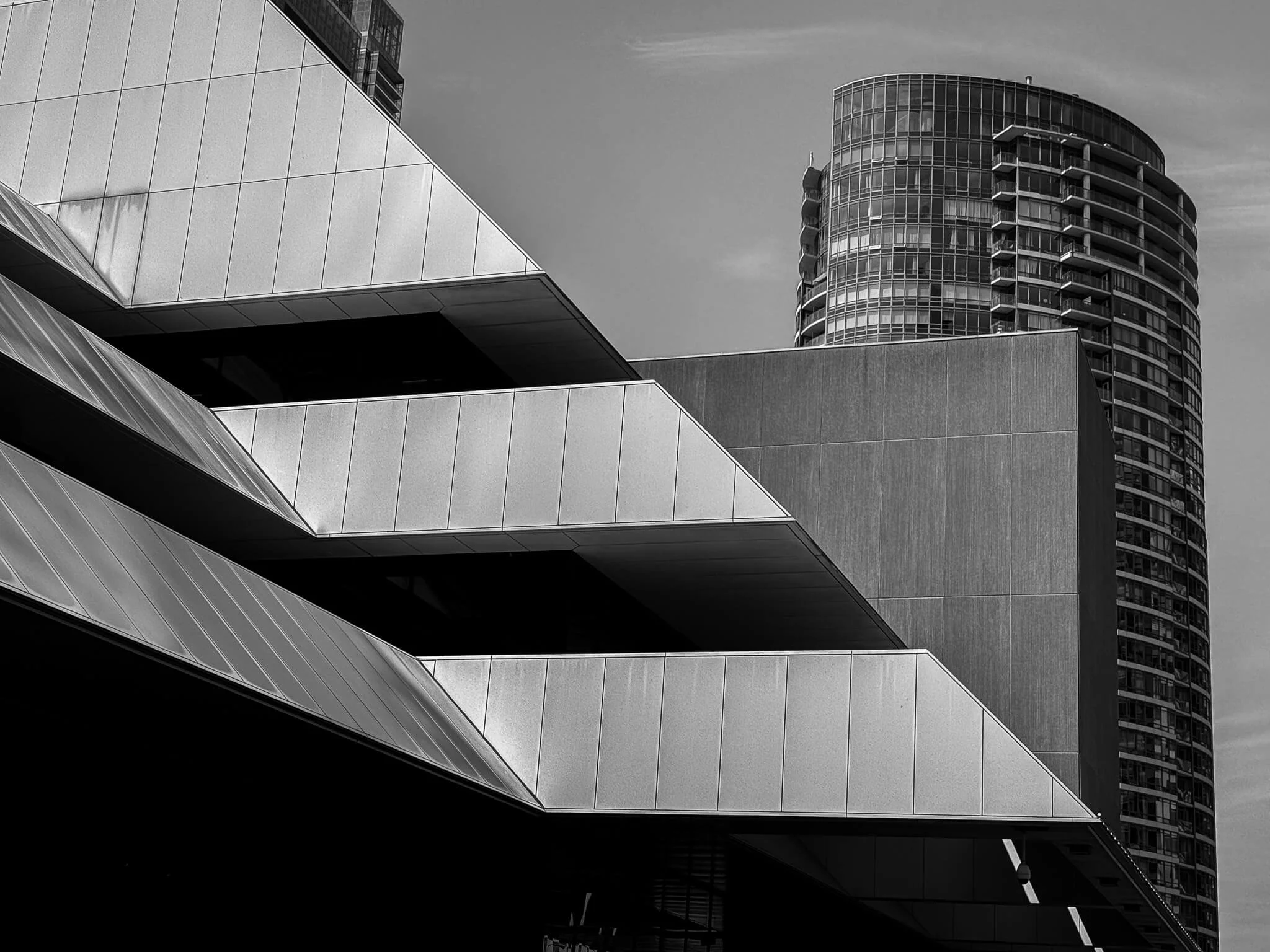 Black and white photo of modern architectural buildings, including a cylindrical high-rise with balconies and a geometric building with angular elements and reflective surfaces.
