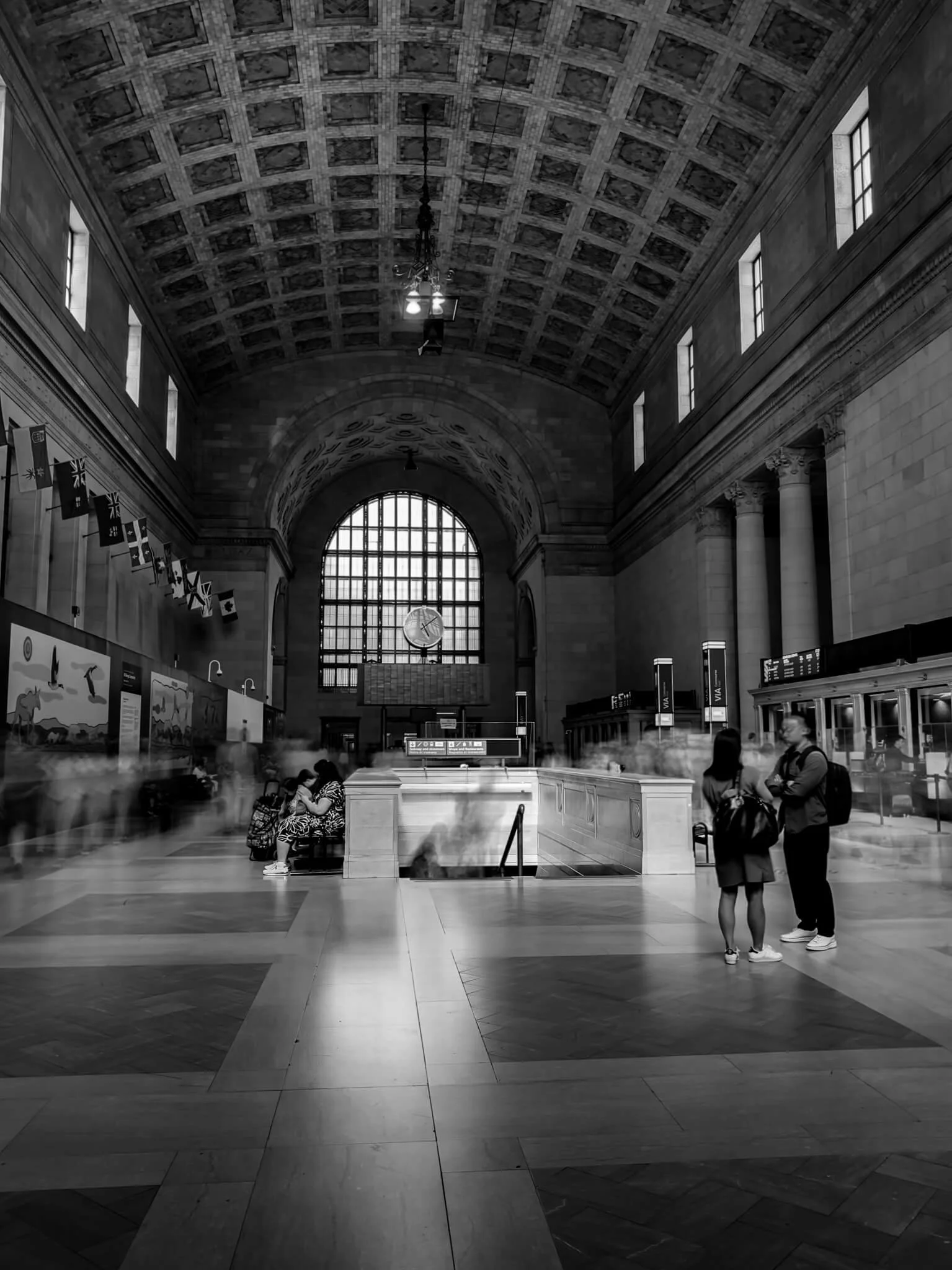 Interior of a grand train station with high arched ceiling, large window, and people waiting and talking.