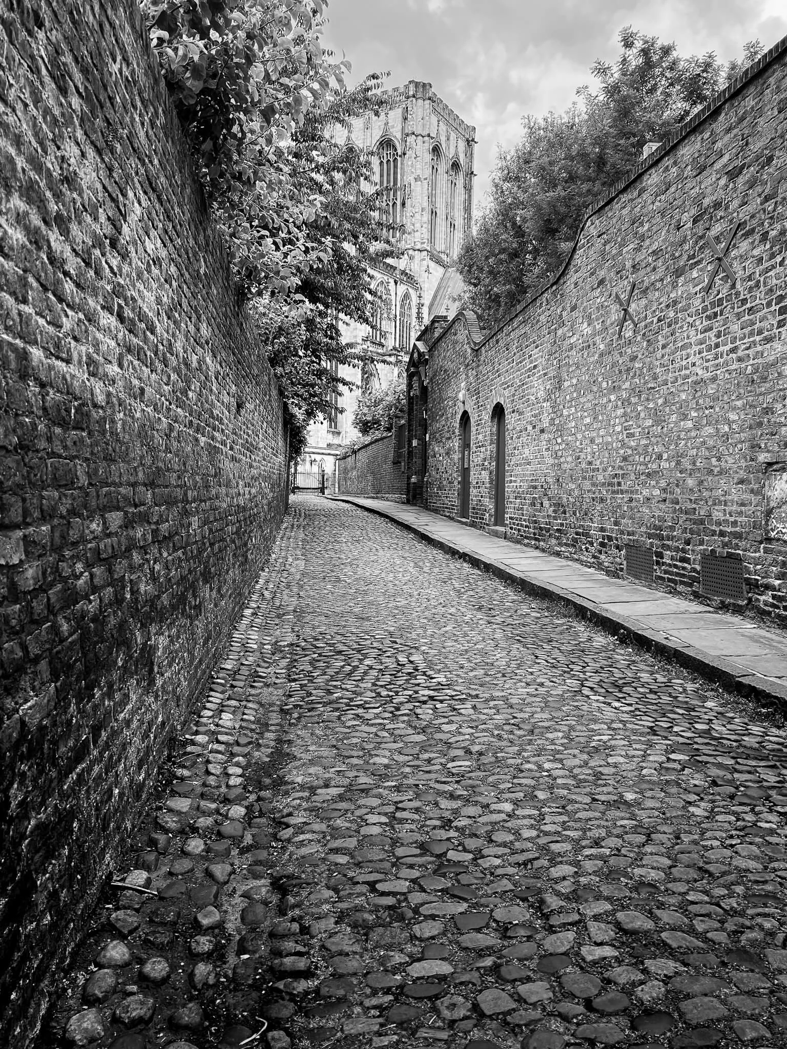 A narrow cobblestone alleyway with brick walls on both sides, leading up to a large Gothic-style cathedral visible in the distance, with trees peeking over the walls.