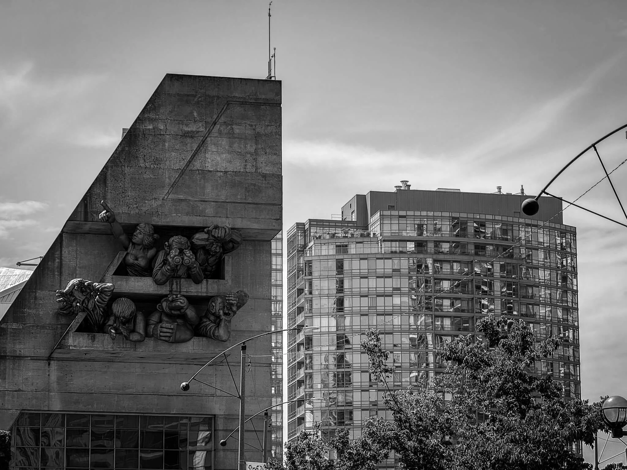Black-and-white photo of a cityscape with modern and historic buildings, including a building with sculptures of workers and a glass office tower, with string lights and trees in the foreground, under a partly cloudy sky.