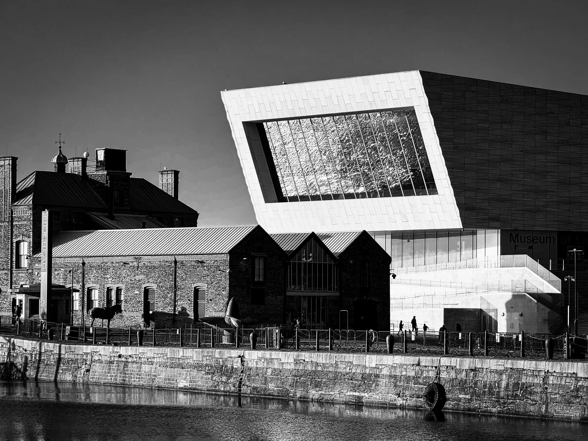 Historical brick buildings next to a modern glass and metal building with a tilted facade, along a waterfront with pedestrians walking and a boat docked in the water.