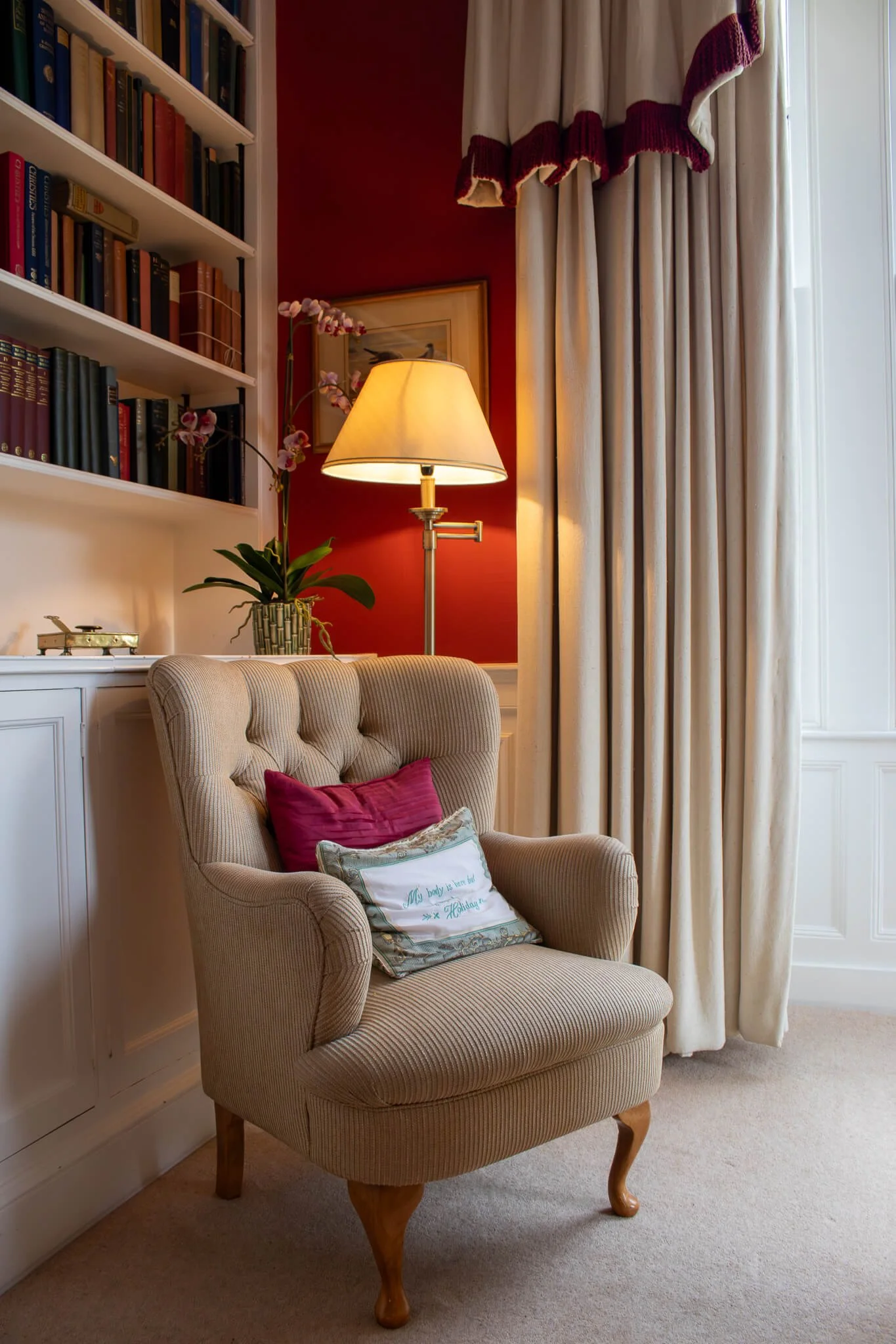 A cozy room with a beige, tufted armchair with wooden legs, decorated with two pillows, one pink and one with embroidery, near a large window with white curtains and maroon valance. A tall floor lamp with a yellow shade stands beside a white bookshelf filled with books, against a red wall with framed artwork and a potted orchid plant on a white cabinet.