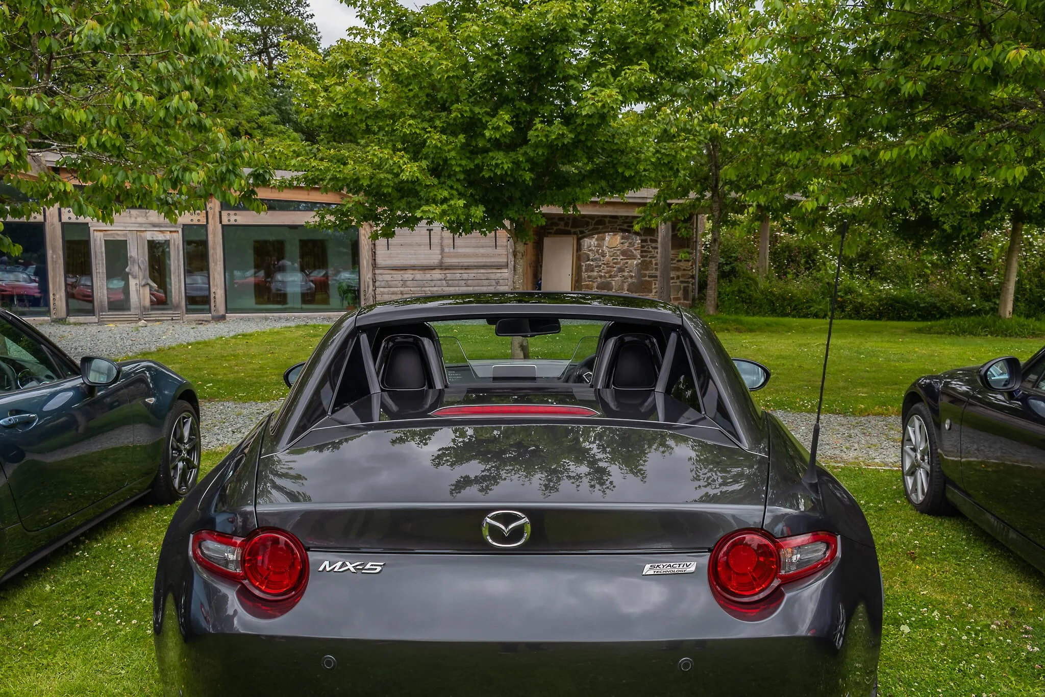 Black Mazda MX-5 convertible sports car parked on grass, with two other cars on either side, under a large leafy tree, in front of a rustic building with glass windows.
