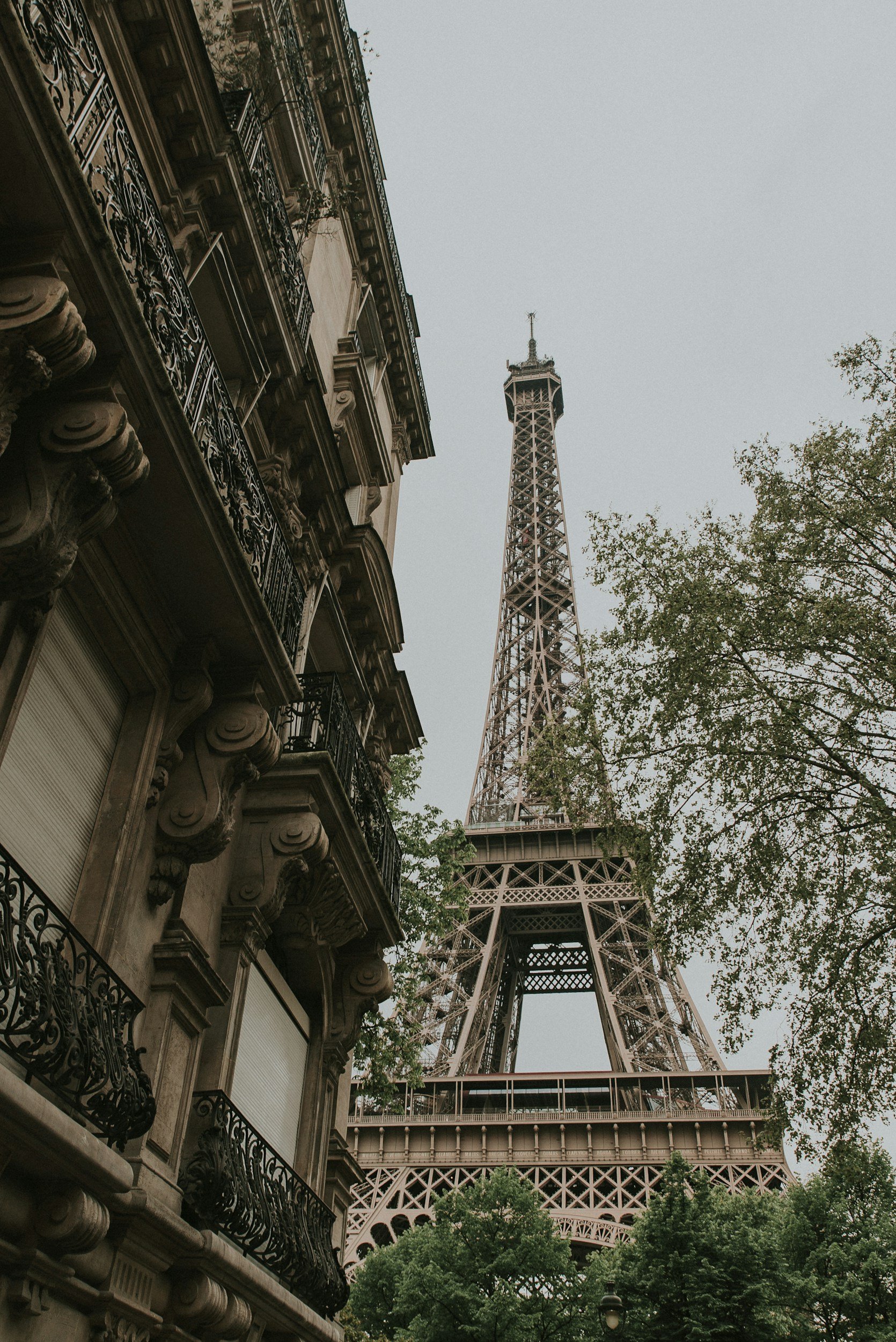 Low-angle view of the Eiffel Tower, with a historic Parisian building and trees nearby on an overcast day.