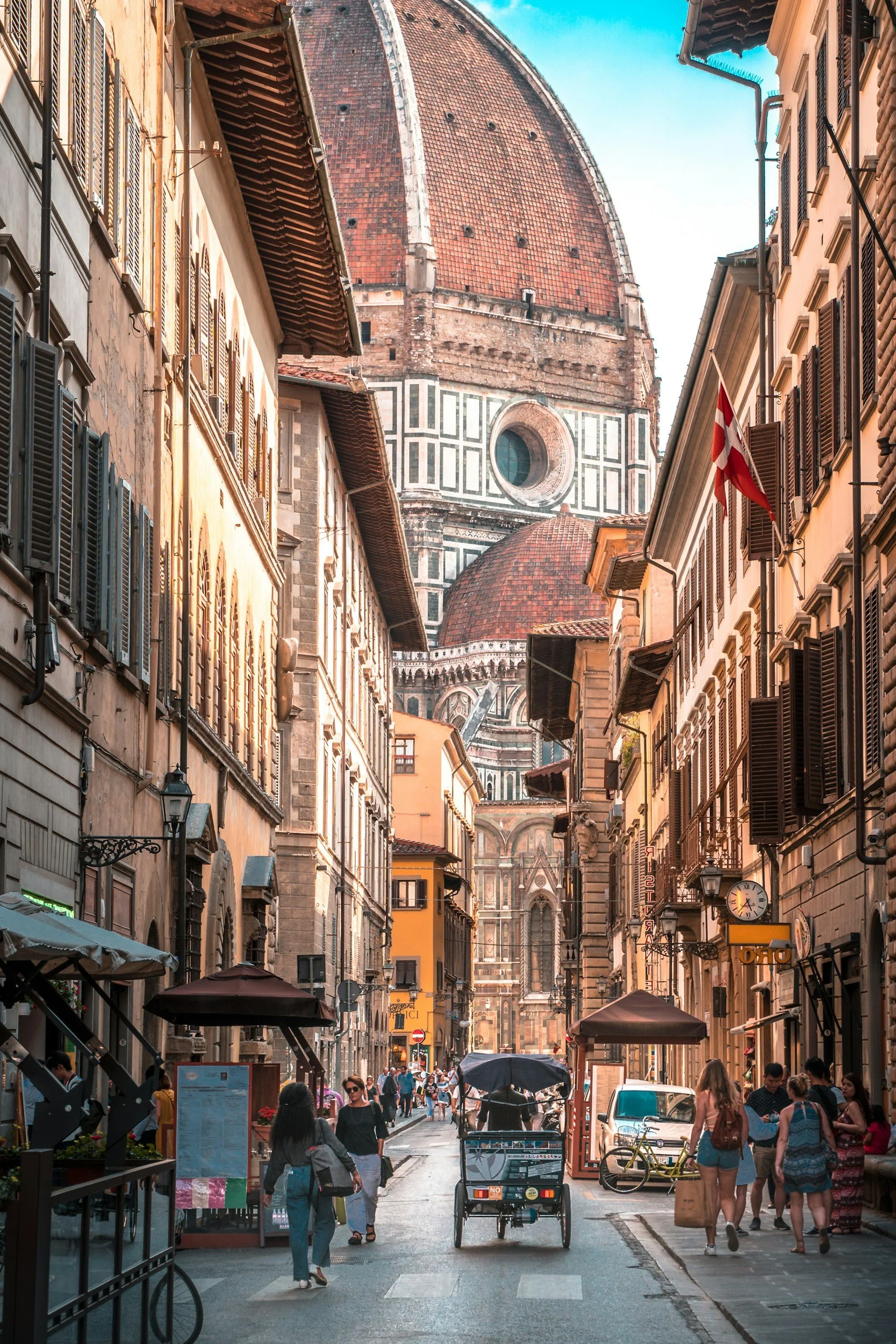 A busy street in Florence, Italy, with tall historic buildings, cafes, and shops lining the street, leading towards the Florence Cathedral with its large red dome in the background.