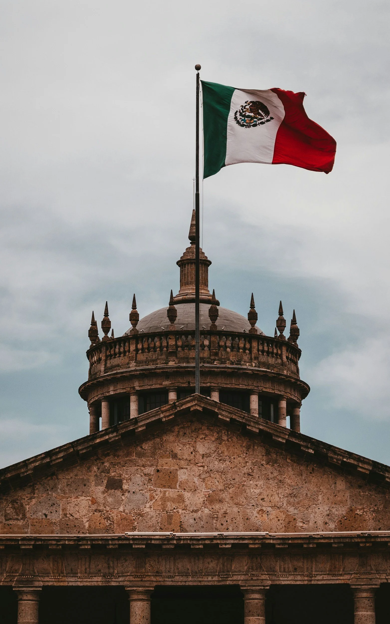Mexican flag flying atop an old stone building with a rounded tower and spiked decoration, under a cloudy sky.