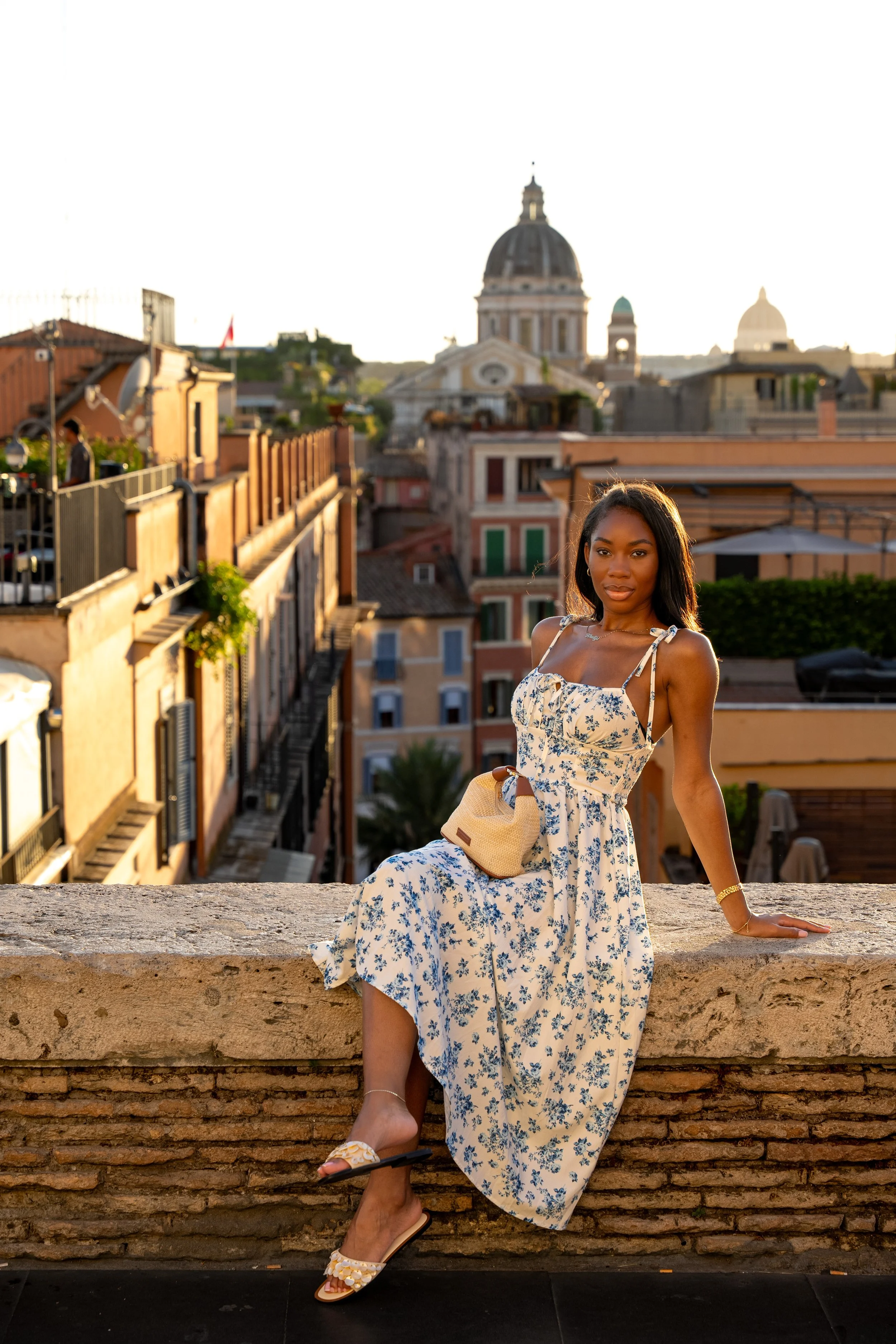 Young woman in a floral dress sitting on a brick ledge with Rome's cityscape and domed churches in the background during sunset.