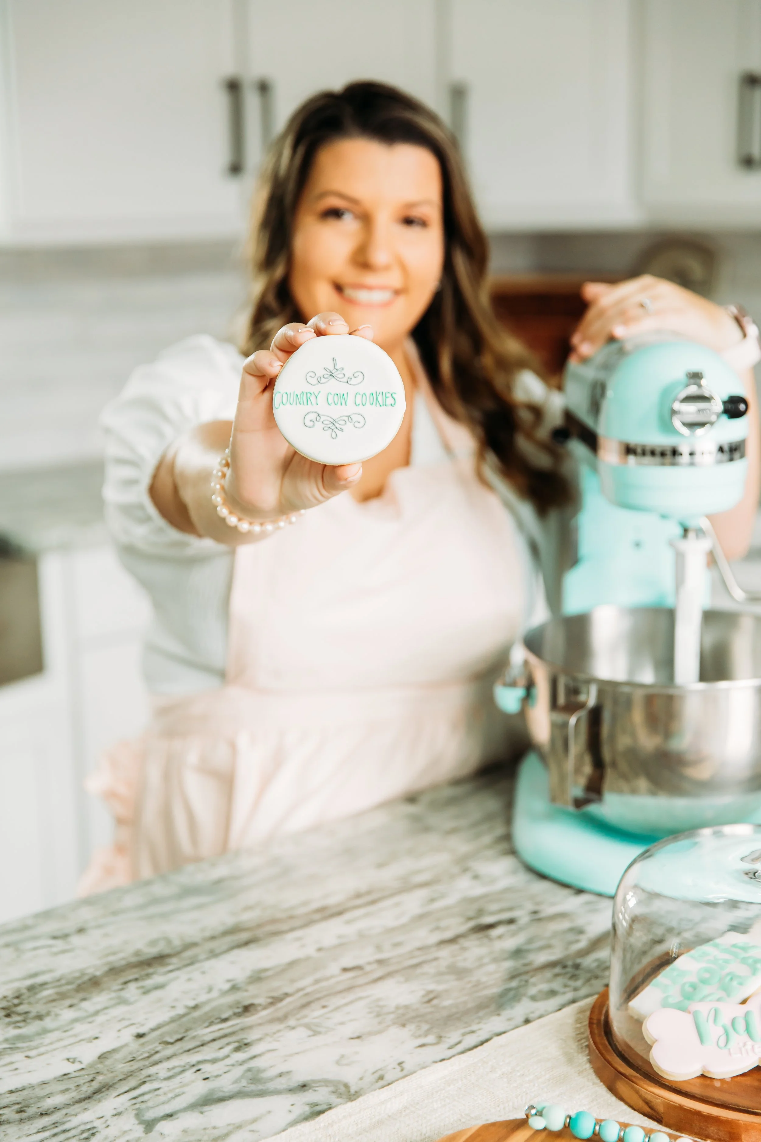 Bakerin a light pink apron holding a cookie with the words 'Country Cow Cookies' written on it, standing in a kitchen with a stand mixer and cookie decorations nearby.