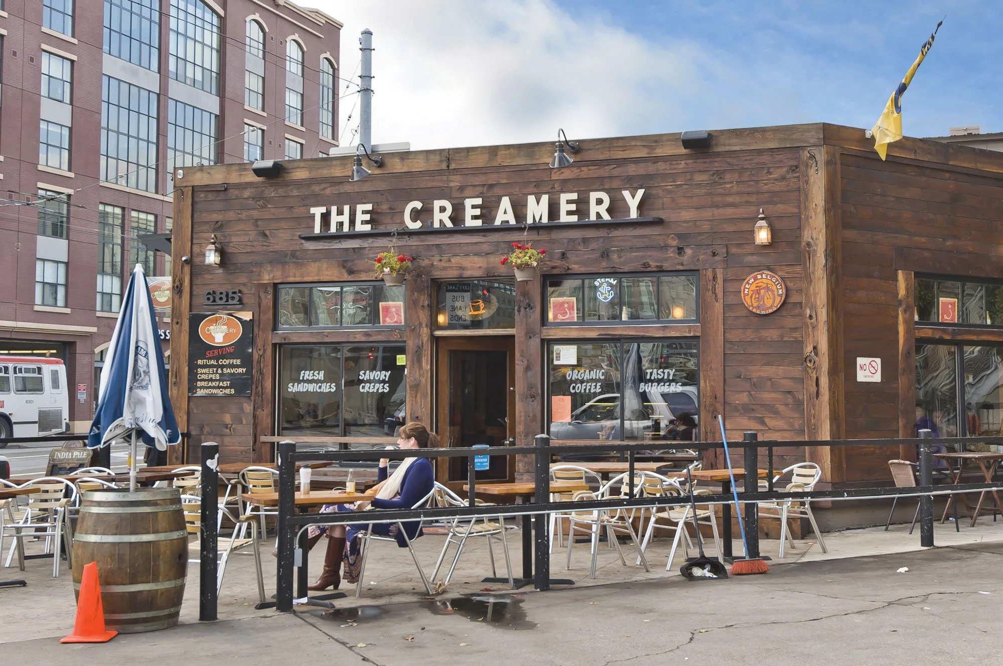 Wooden exterior of The Creamery restaurant with outdoor seating, a woman sitting at a table, and a cityscape in the background.