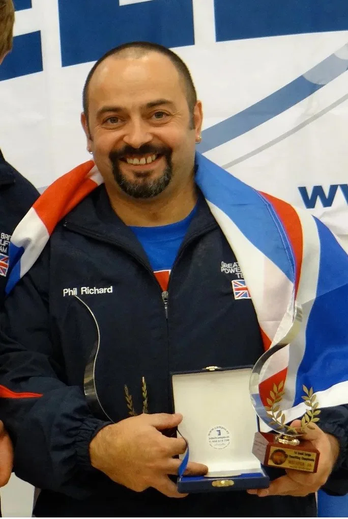 Smiling man with a goatee and short hair holding a trophy and award in front of a blue and white background, wearing a dark jacket with a name patch that reads Phil Richard.