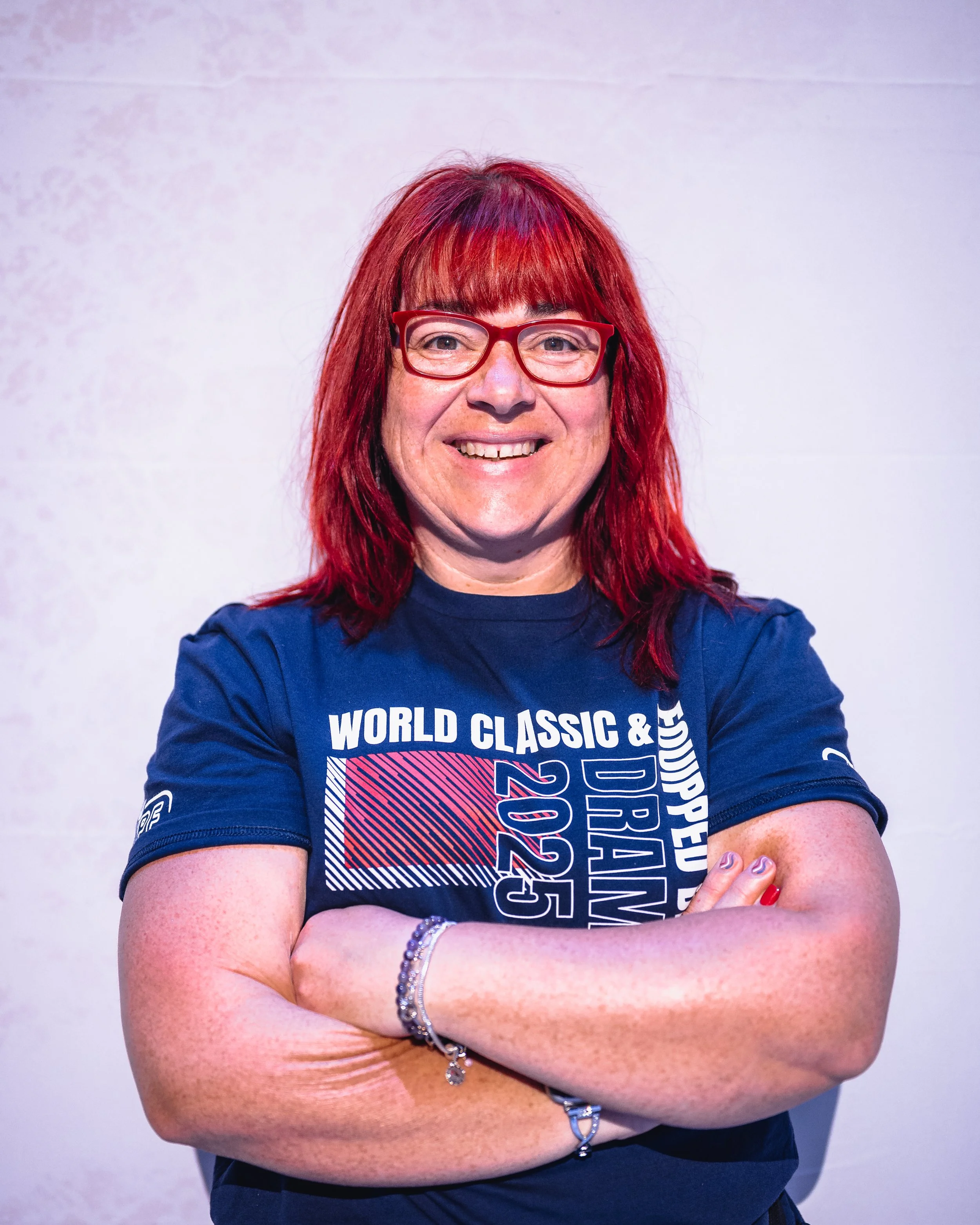 Woman with red hair and glasses smiling with arms crossed, wearing a dark blue t-shirt with printed text and graphics, standing against a plain light-colored background.