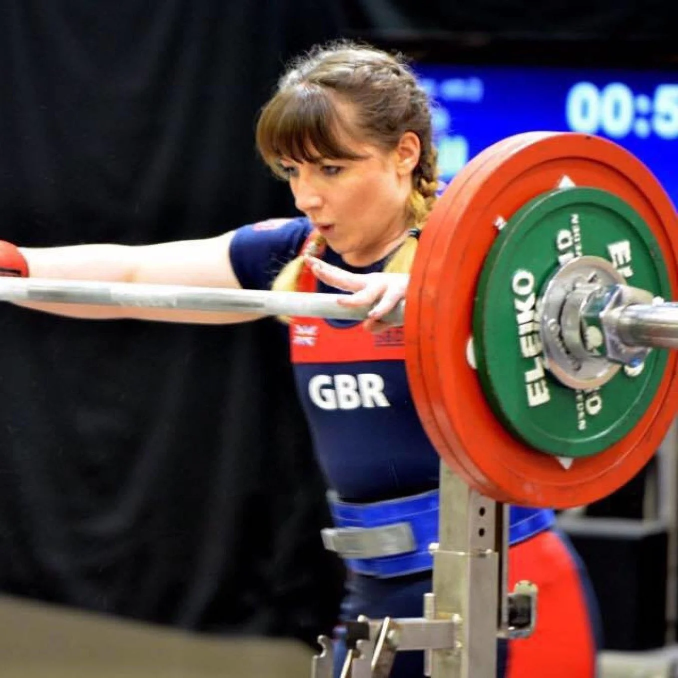 A female weightlifter with braided hair is focusing while lifting a barbell with orange and green weights marked 'ELEIKO' during a competition, wearing a black and blue outfit with 'GBR' on it.