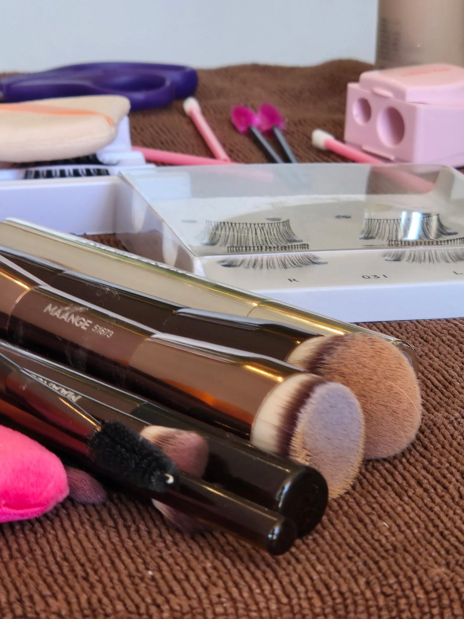 A collection of makeup brushes, scissors, a pink plastic container, and a pair of pink tongs on a brown textured surface.