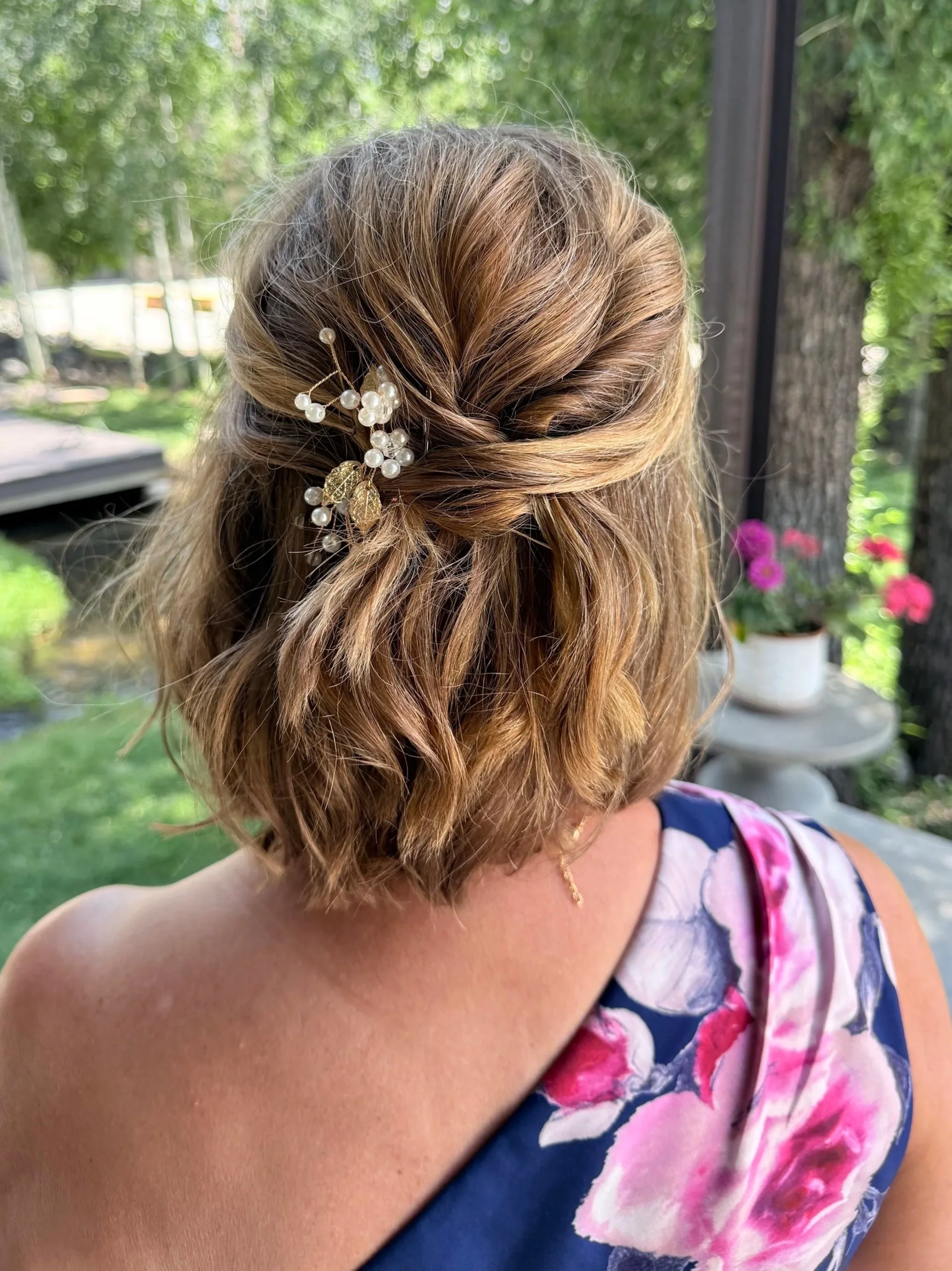A woman with shoulder-length, wavy, light brown hair styled with a pearl hair accessory featuring gold leaves. She is wearing a sleeveless dress with a floral pattern in pink, purple, and white, and a gold necklace. The background shows greenery and blooming pink flowers.