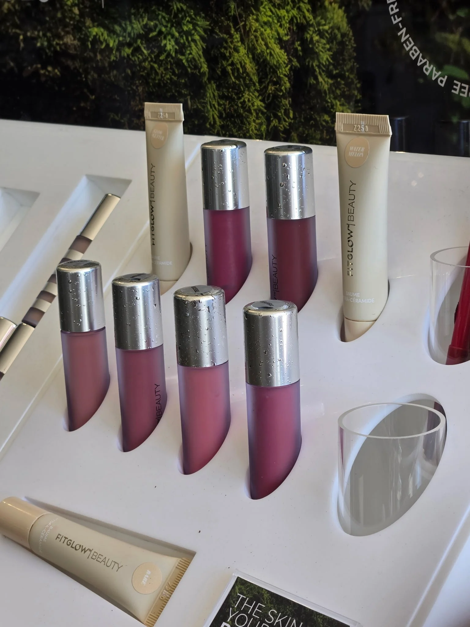 Display of skincare products including tubes labeled 'FITGLOW BEAUTY' and bottles with silver caps, arranged on a white surface with a mossy green background.