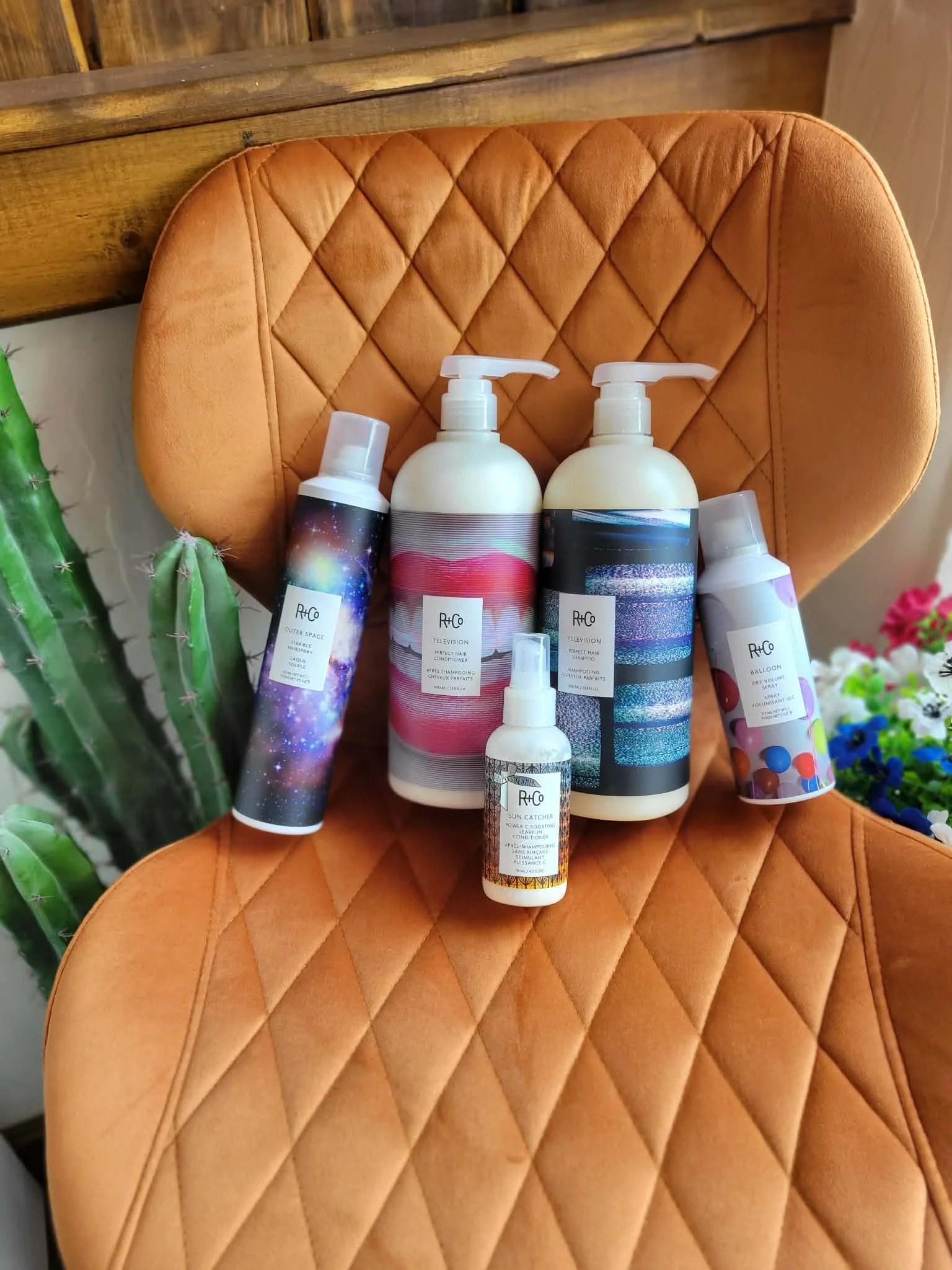 Various bottles of hair care products placed on a tan, quilted chair with a cactus plant on the side.