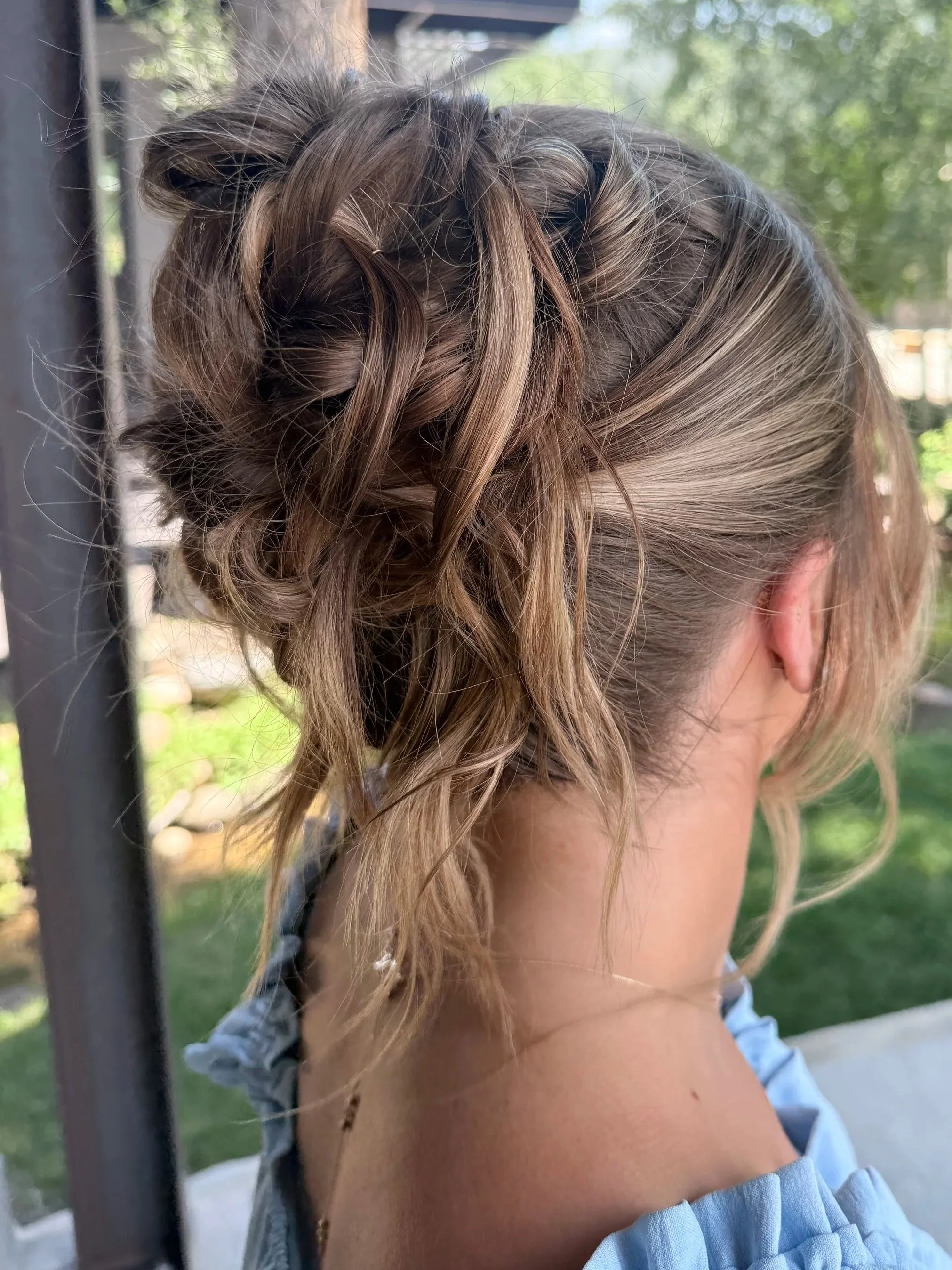 Close-up of a woman with curly, light brown hair styled in a braided updo, wearing a light blue off-the-shoulder top, outdoors with greenery in the background.