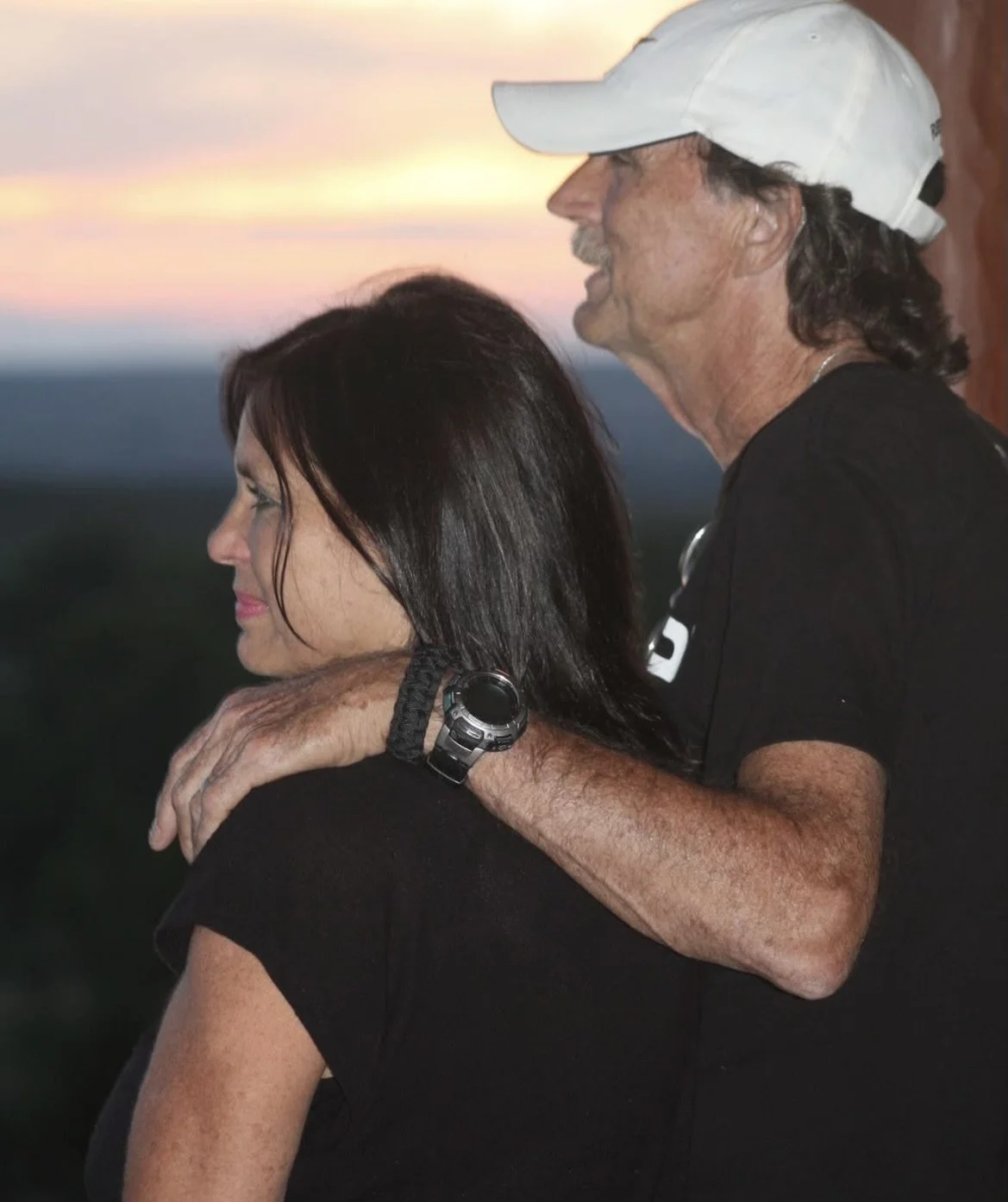 Donna and Bill Wilting, both wearing black shirts, watching a sunset outdoors. The man is wearing a white cap.