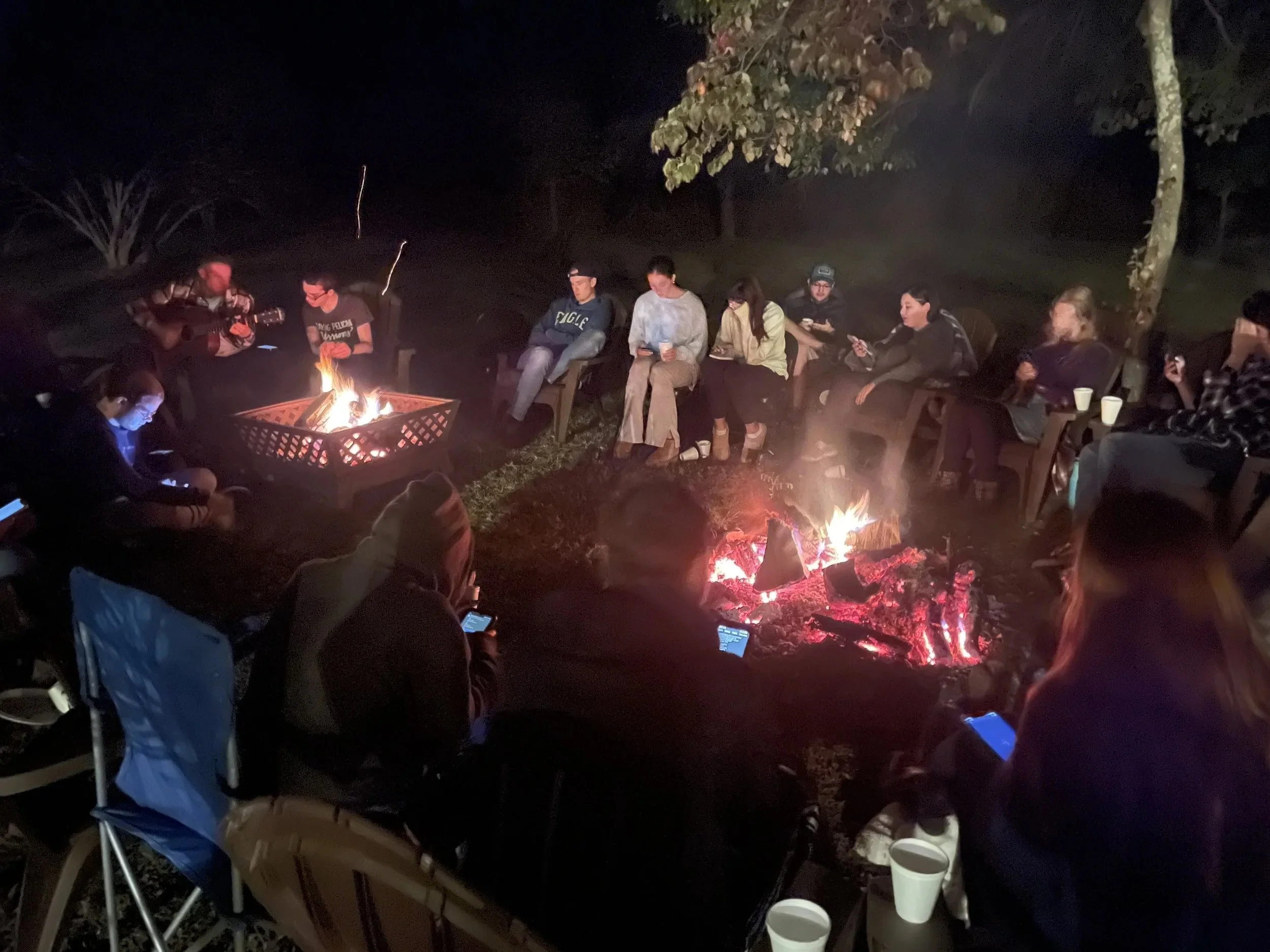 A group relaxes fireside around two campfires at night, some chatting while others look at their phones.