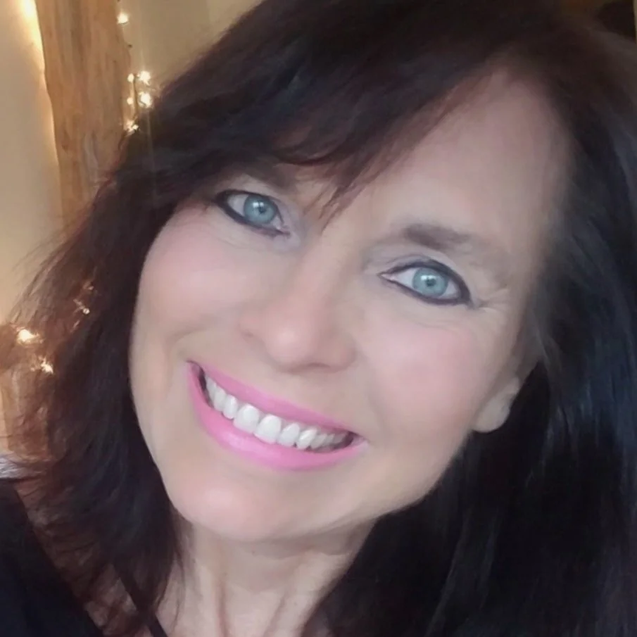 Donna Wilting, a smiling woman with bright blue eyes and dark hair, poses indoors with soft lighting and string lights behind her.