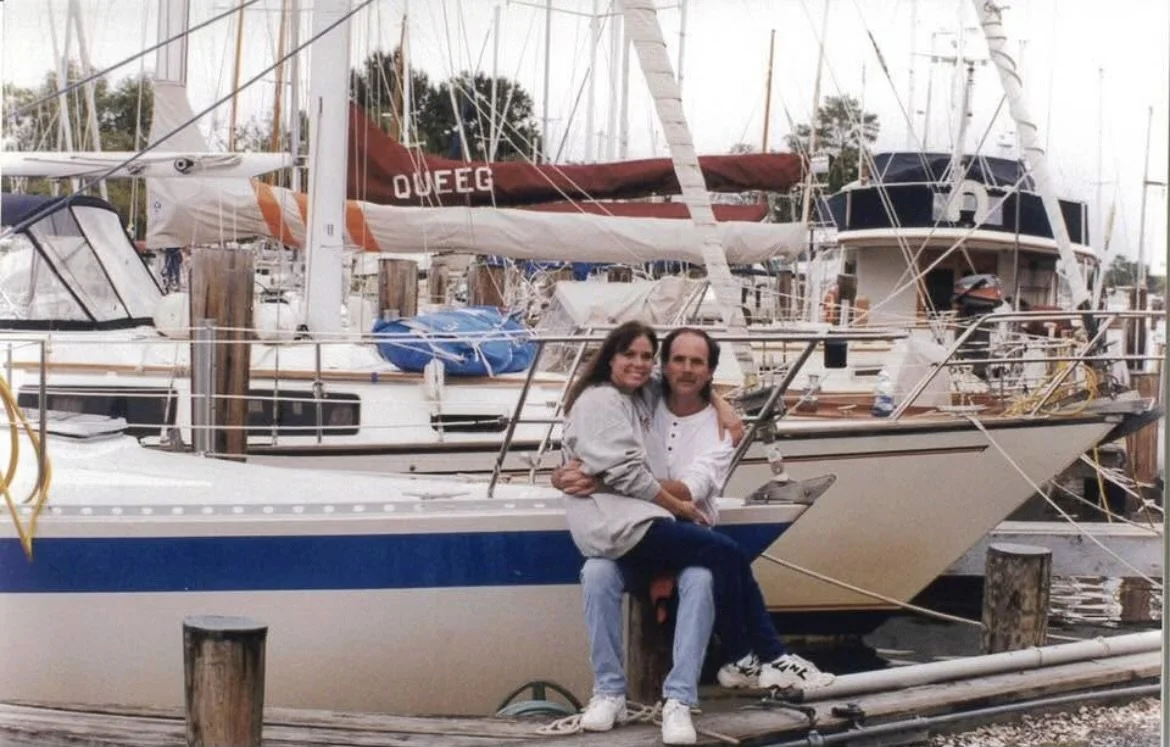 Donna and Bill Wilting, DYM founders sit together on a dock in front of sailboats at a marina, smiling and posing for the photo.