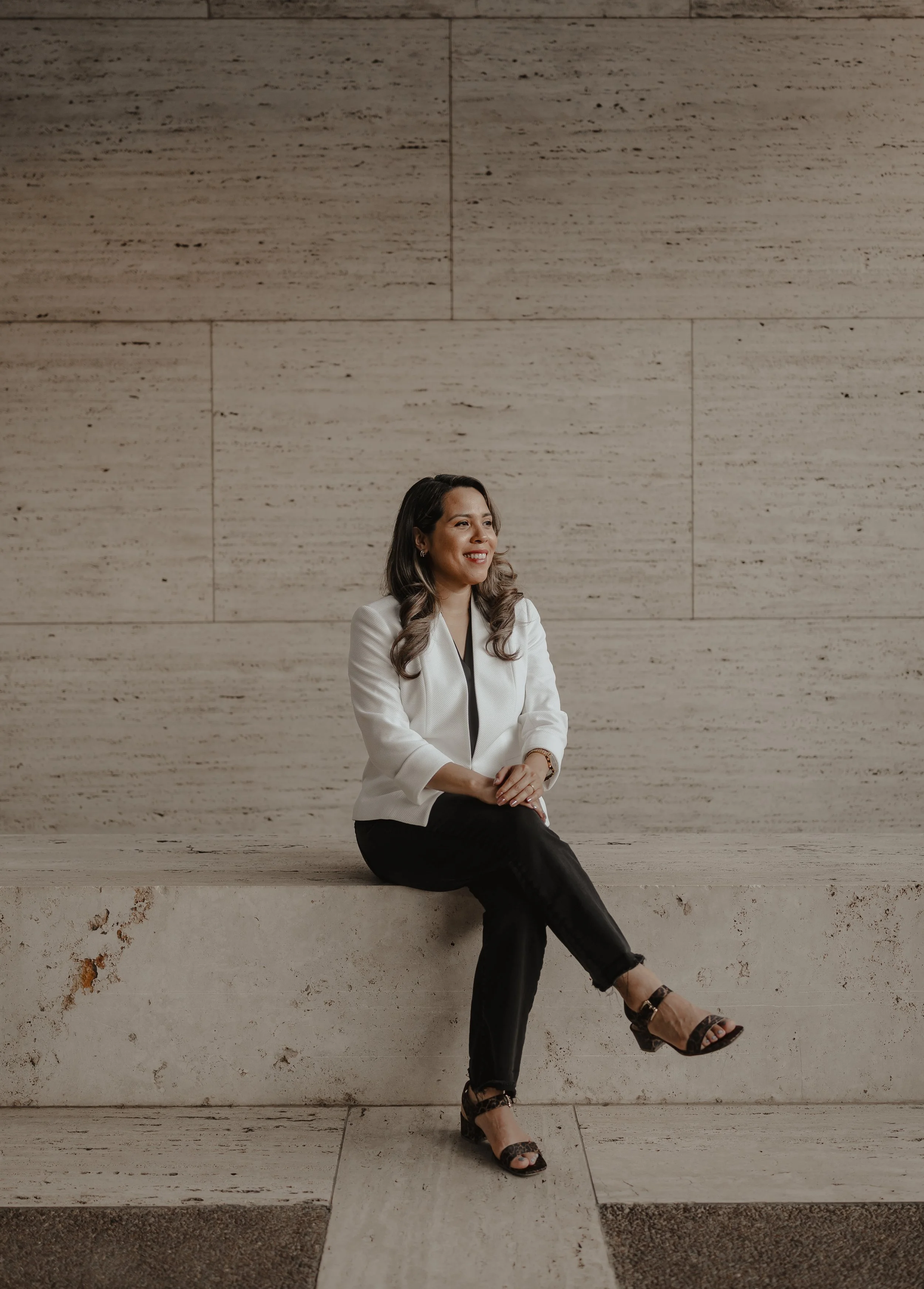 A woman with long wavy brown hair sits on a beige stone ledge against a matching beige stone wall. She is smiling, wearing a white blazer, black pants, and black high-heeled sandals.