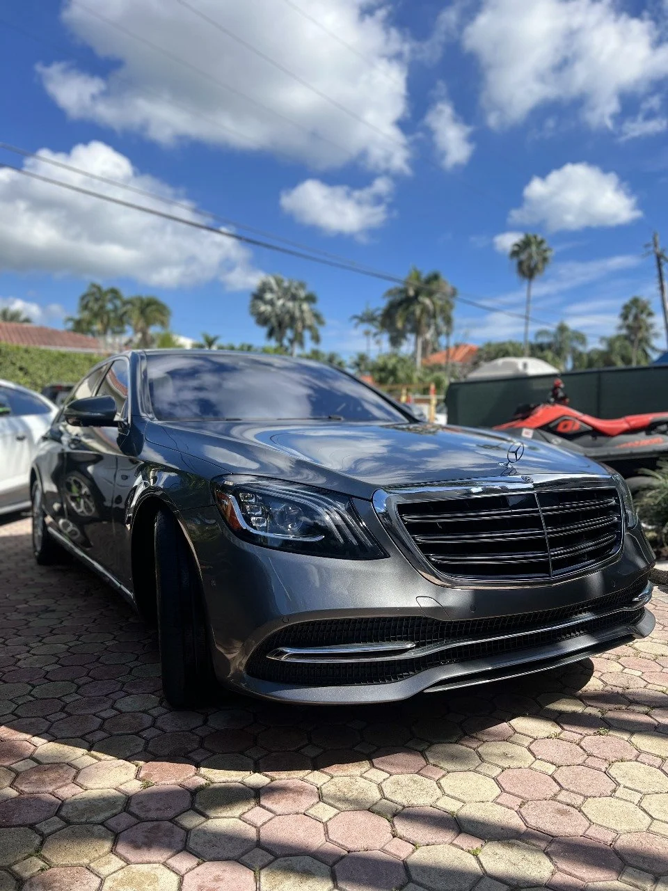 A black Mercedes-Benz sedan parked on a brick-paved lot on a sunny day with a partly cloudy sky, palm trees, and other vehicles in the background.