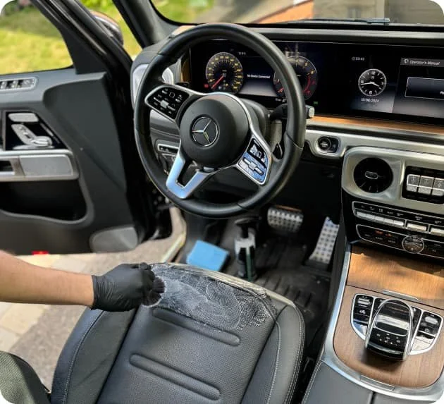 Interior of a Mercedes-Benz vehicle focusing on the steering wheel, dashboard, and center console. A person wearing a black glove is wiping the leather armrest of the driver's seat with a cloth.