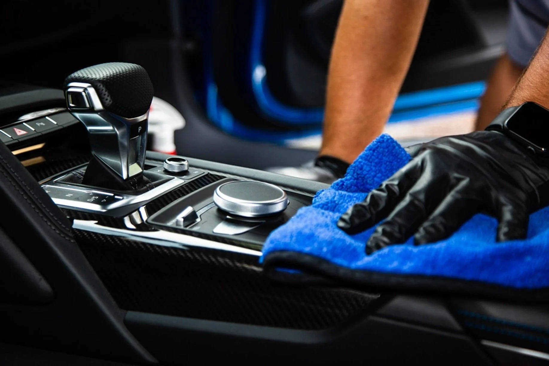 Close-up of a car's center console being cleaned with a blue microfiber cloth. The gear shifter with a textured black knob is visible, along with a circular control knob and other controls. A person's hand wearing a black glove is seen holding the cloth, and part of their arm and a smartwatch are visible.