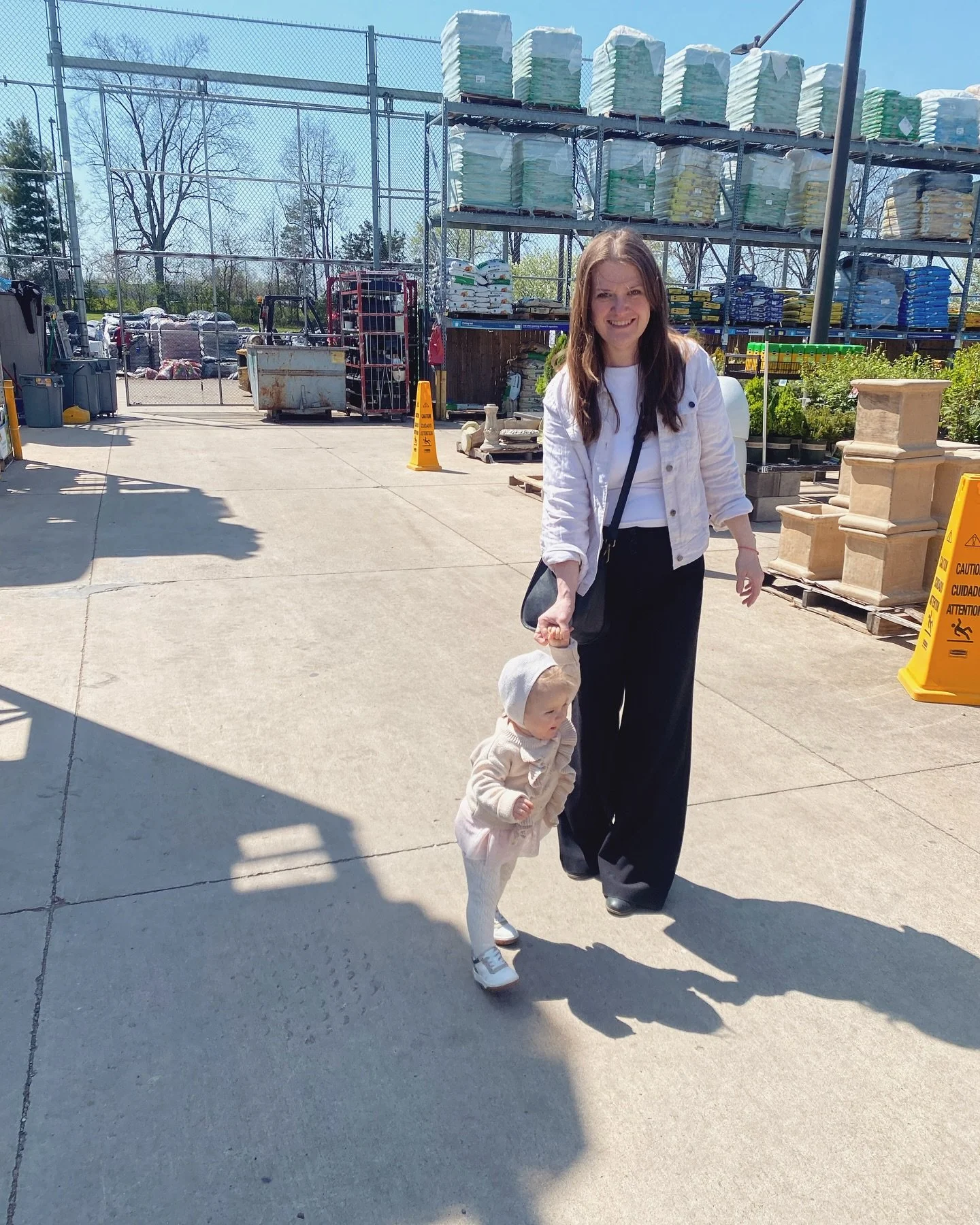 A woman holding a child's hand walking outdoors in a store parking lot on a sunny day. The woman is smiling, wearing a white jacket and black pants, and the child is dressed in light-colored clothes and a hat. Behind them are storage shelves with gardening supplies and a yellow caution sign on the pavement.