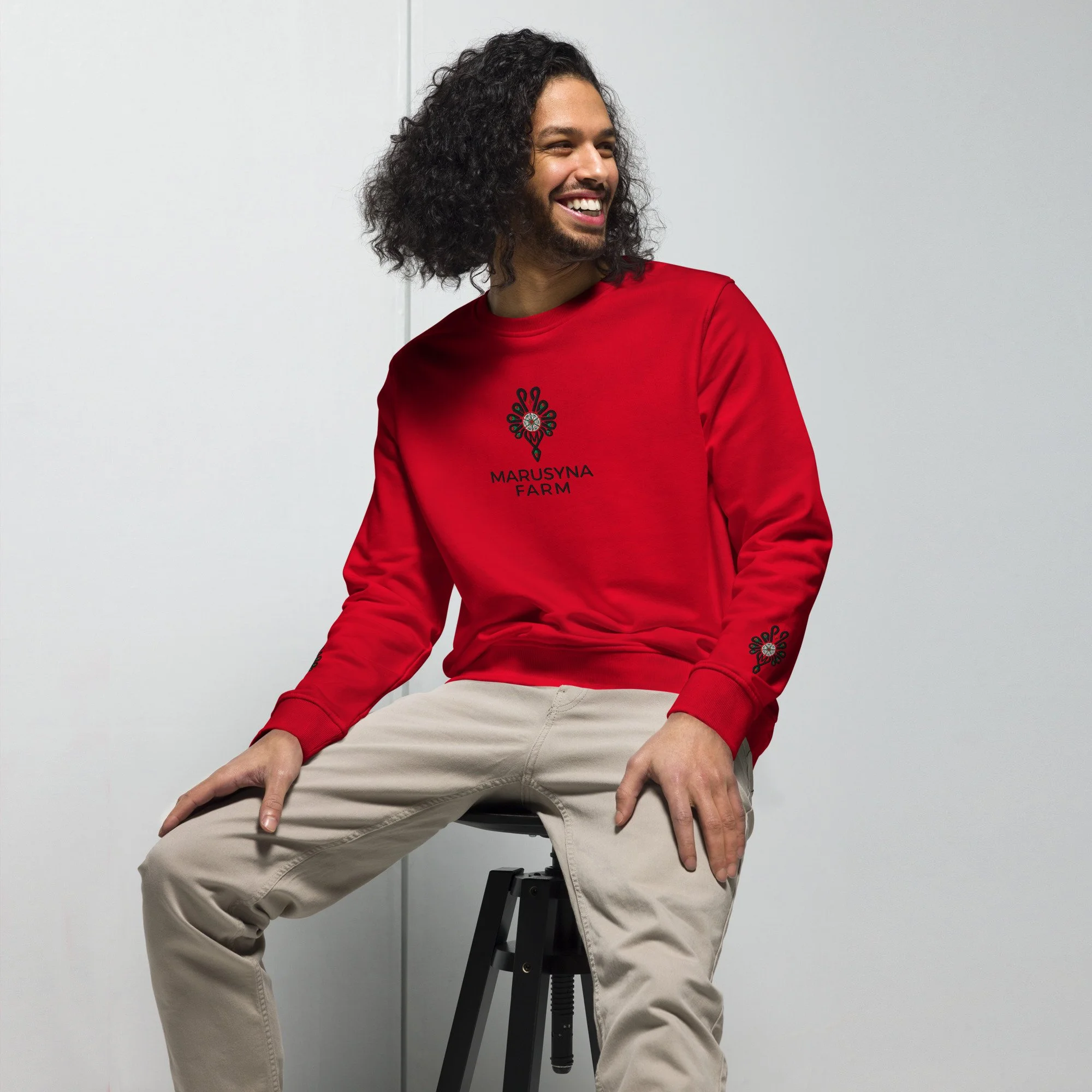 Man with curly dark hair and beard wearing a red Marusyna Farm sweatshirt, sitting on a stool with beige pants, smiling, against a plain light gray wall.