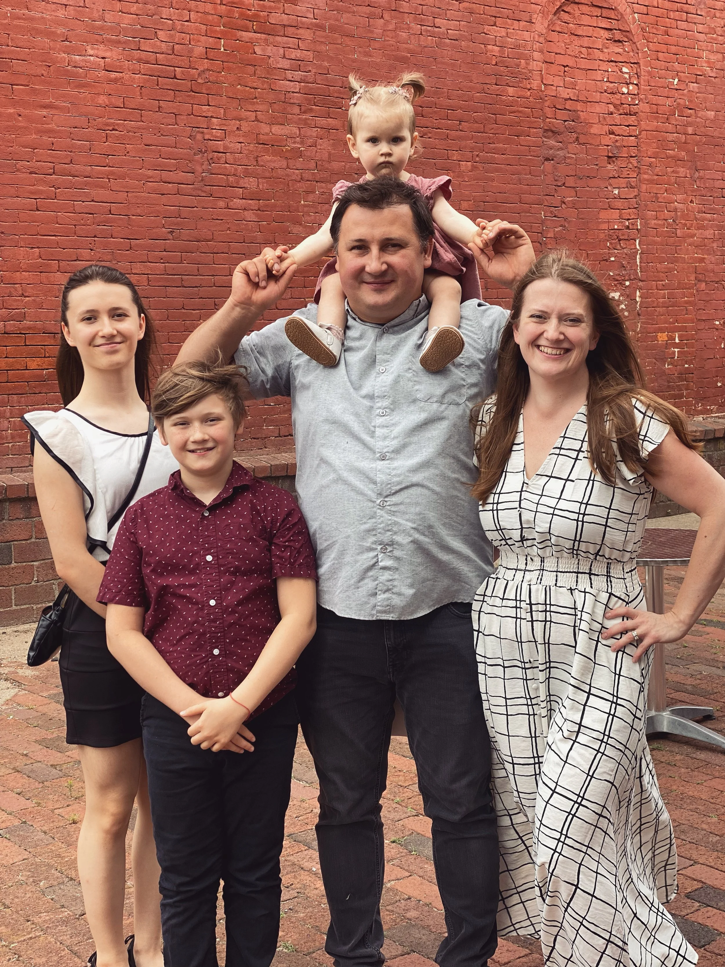 A group of five people, including a man, a woman, two children, and a toddler sitting on the man's shoulders, standing outdoors against a red brick wall. They are smiling and appear happy.