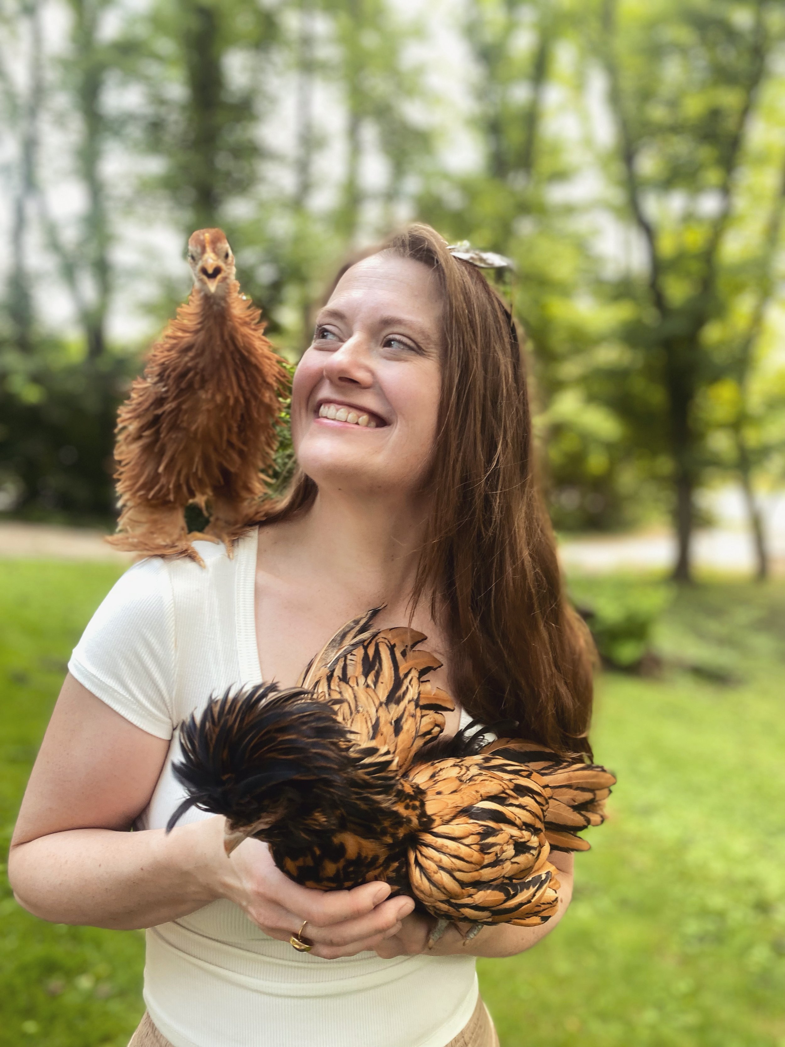 A woman with long brown hair holding a polish chicken, outdoors in a forested area with green trees and grass.